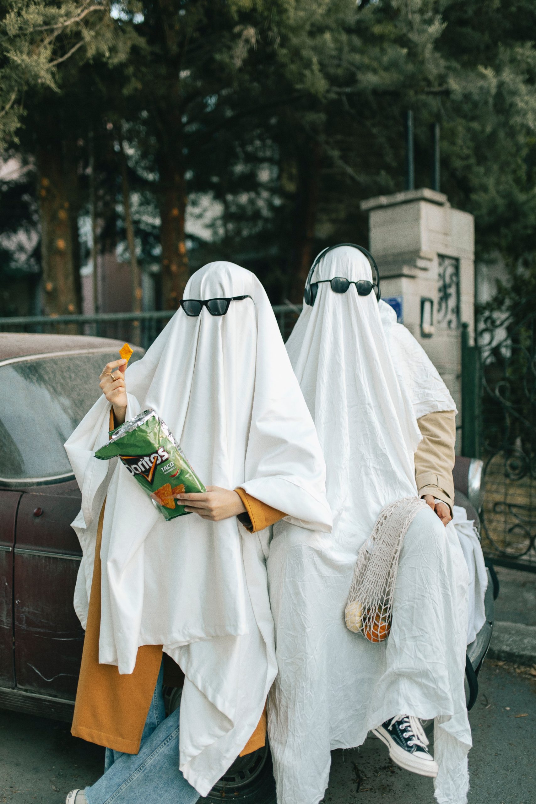 Two people in ghost costumes with sunglasses sit humorously on a car, enjoying snacks.