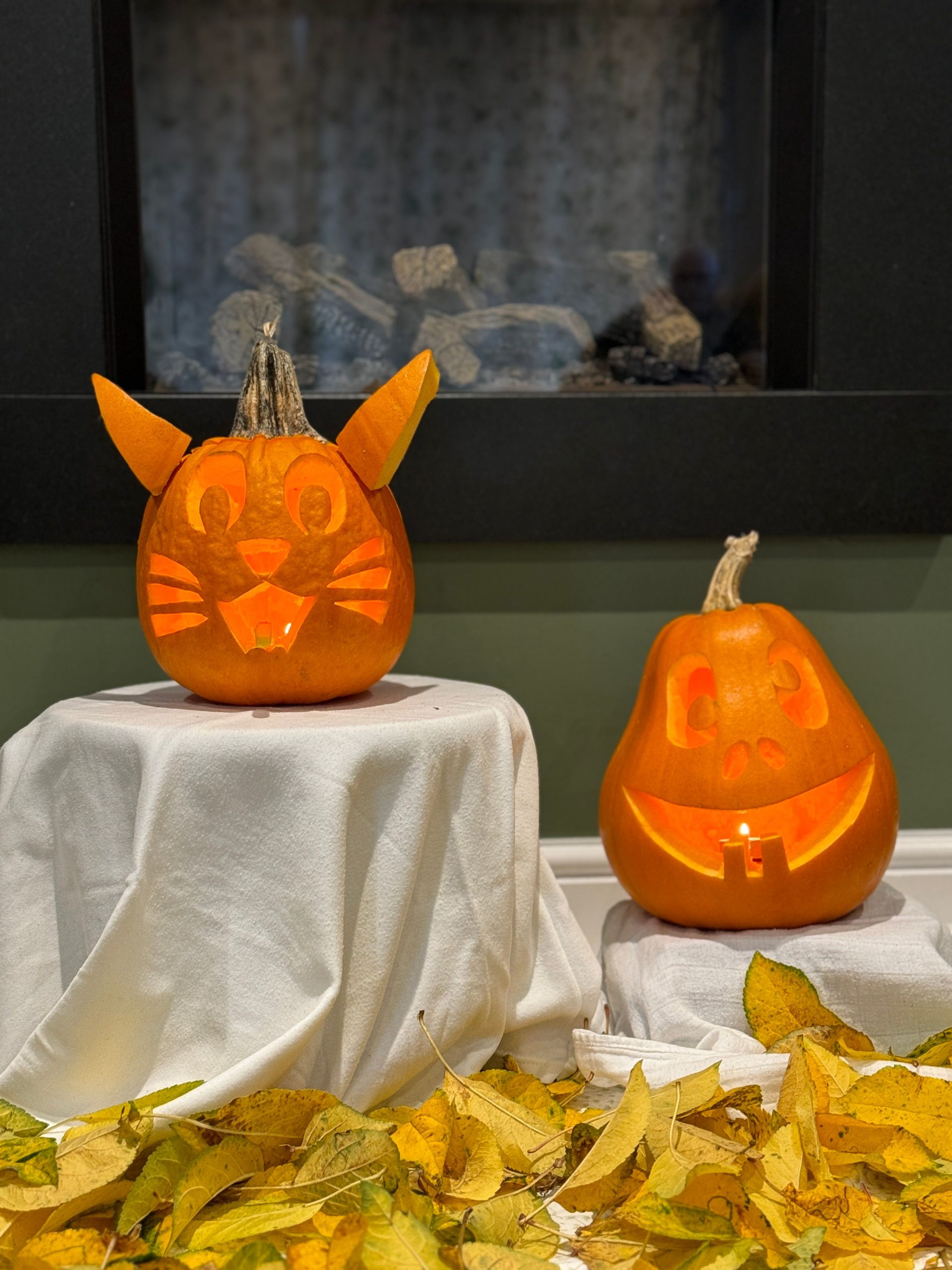 Two uniquely carved pumpkins with autumn leaves in a cozy indoor setting.