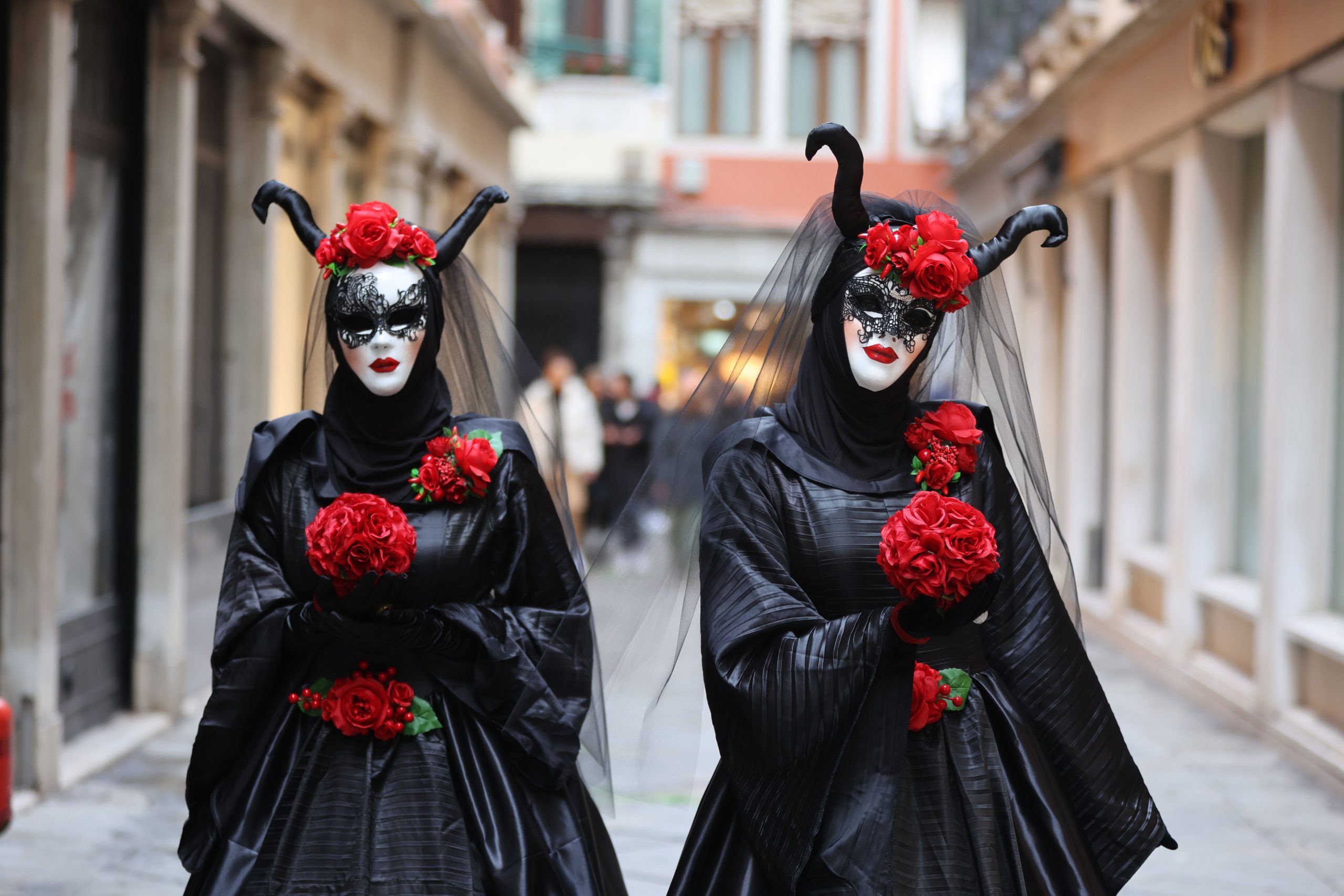 Two women in elaborate black and red costumes with horns and masks on a city street.
