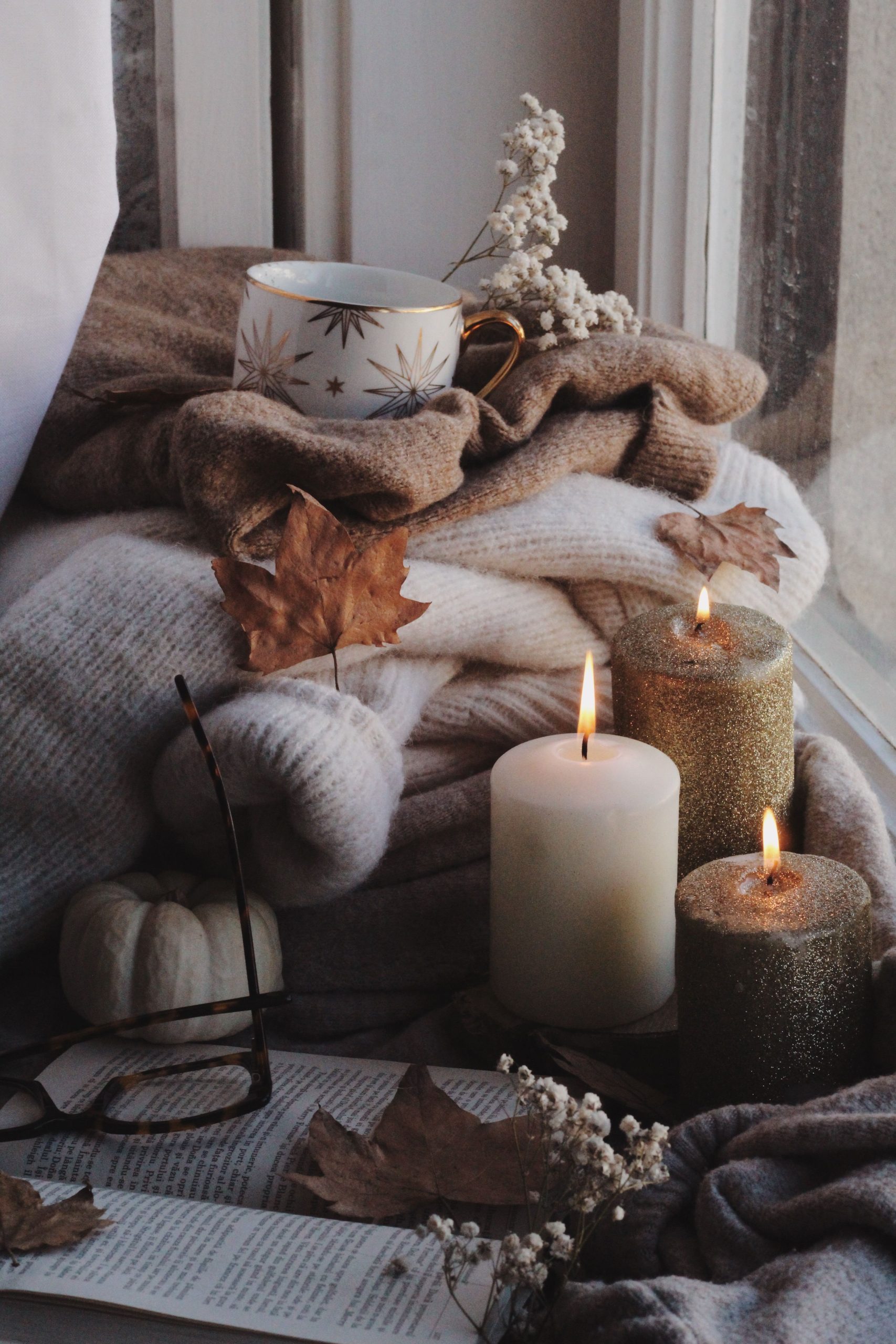 Warm autumn scene with candles, sweaters, and dry leaves by the window, creating a cozy ambiance.