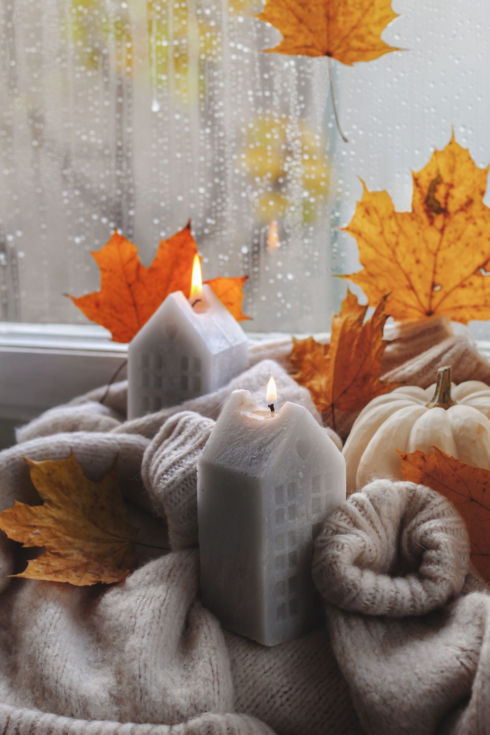 Warm autumn setting with candles, sweater, and leaves by a window, capturing fall's essence.
