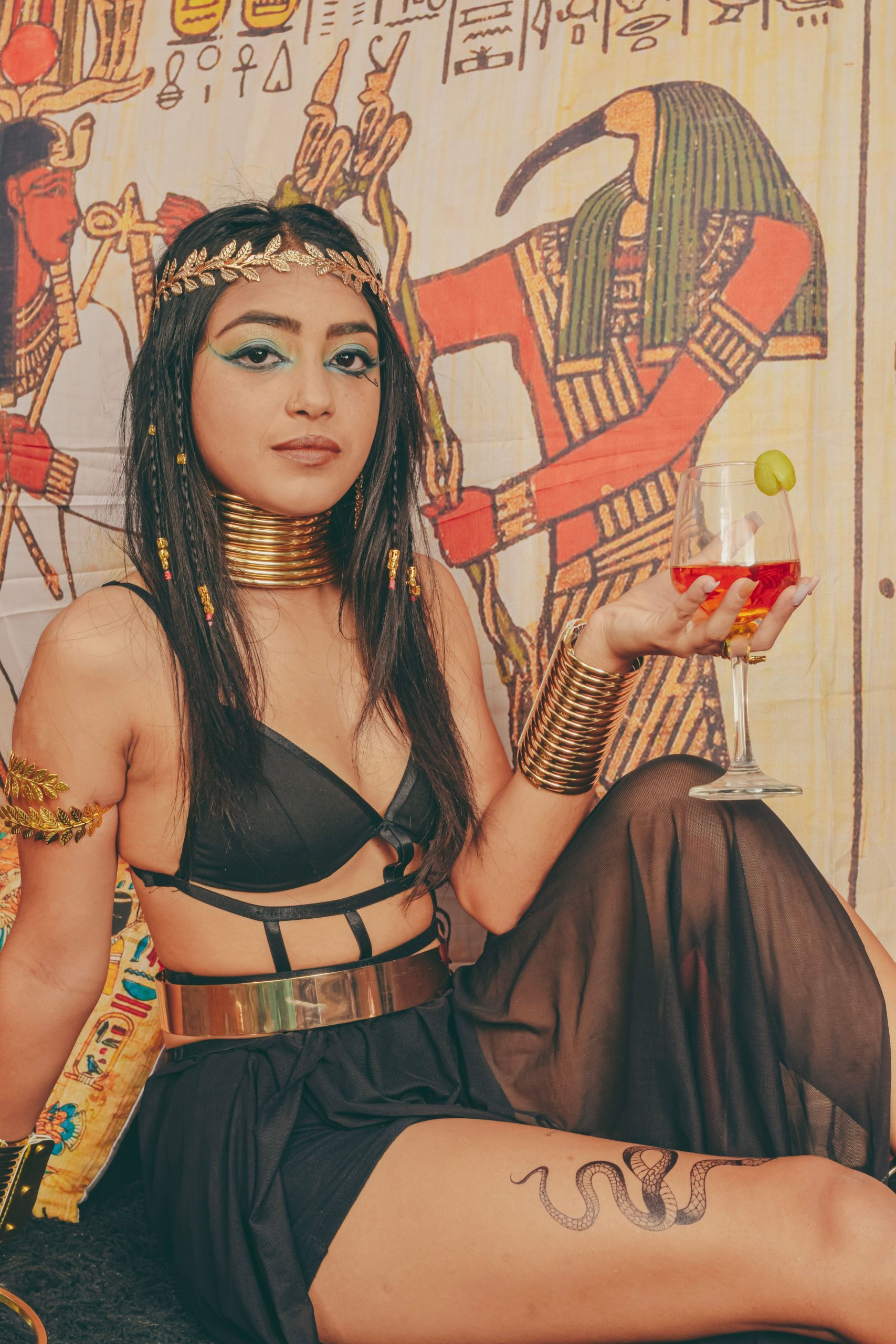 Woman in Egyptian-themed attire with gold accessories holding a cocktail, inspired by ancient art.