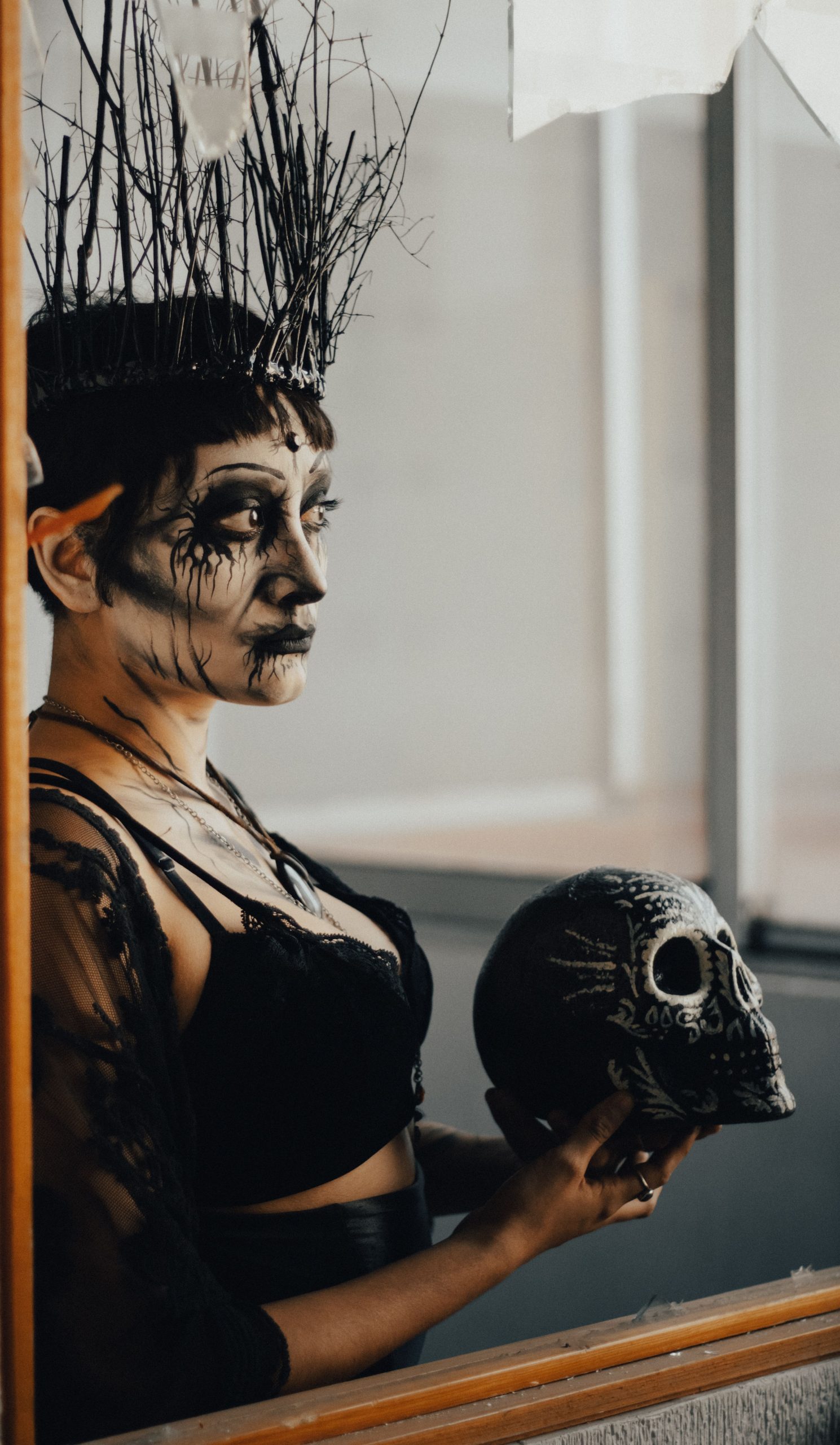 Woman in gothic attire holding a skull with dramatic makeup and headpiece, reflecting in a mirror.