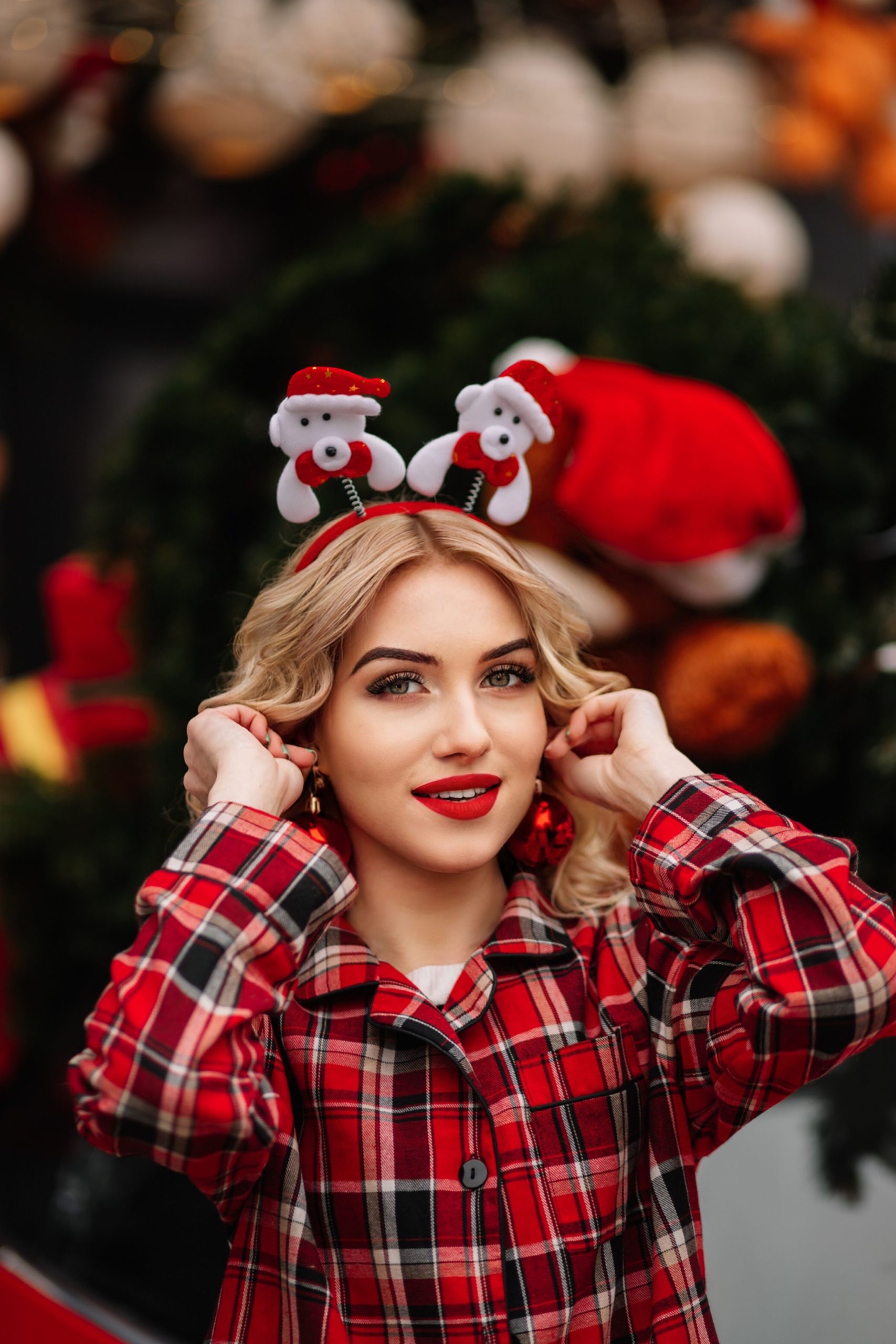 Woman in red plaid pajamas and Santa headband celebrating Christmas indoors with a festive background.