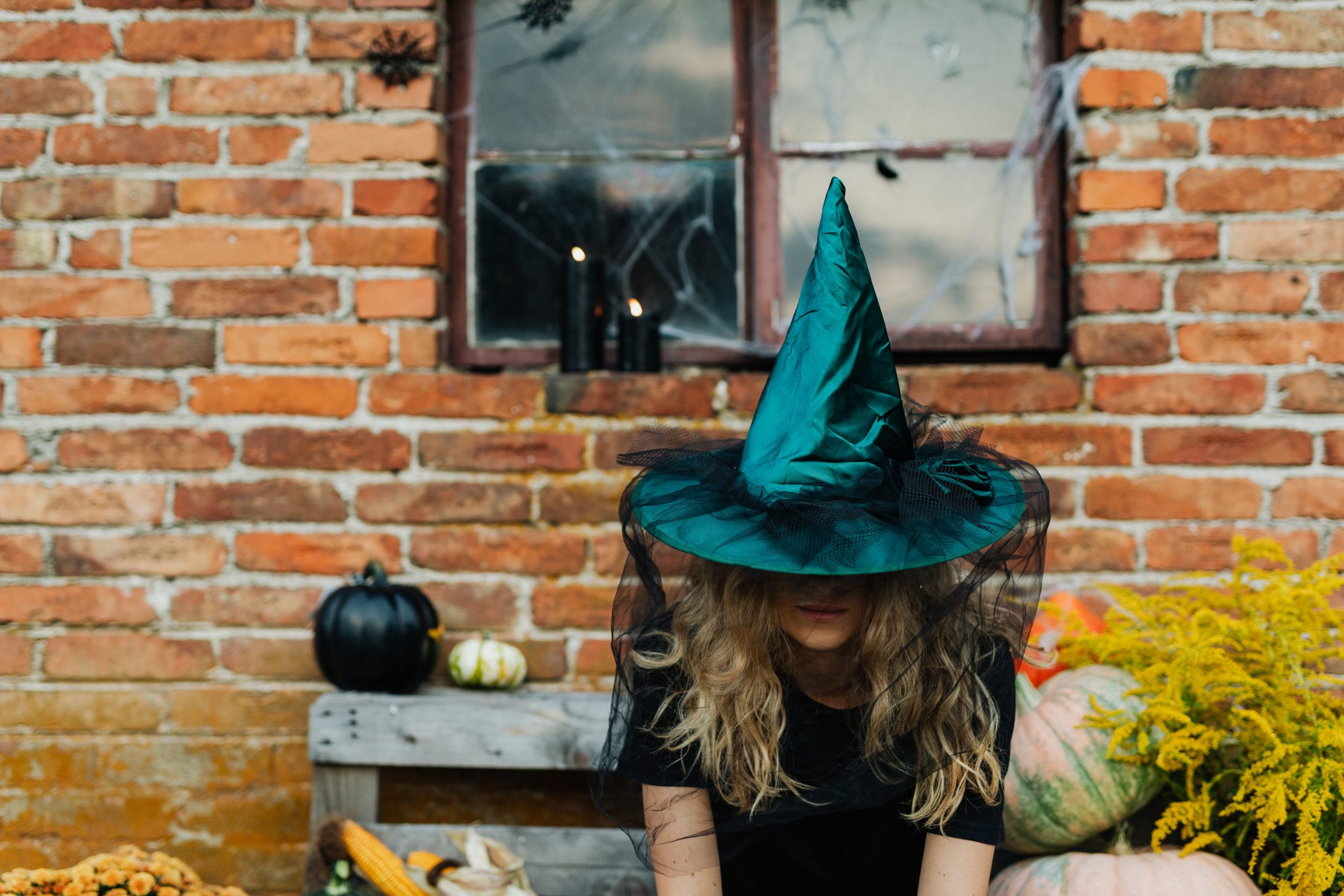 Woman in witch hat sitting outdoors against brick wall with pumpkins.