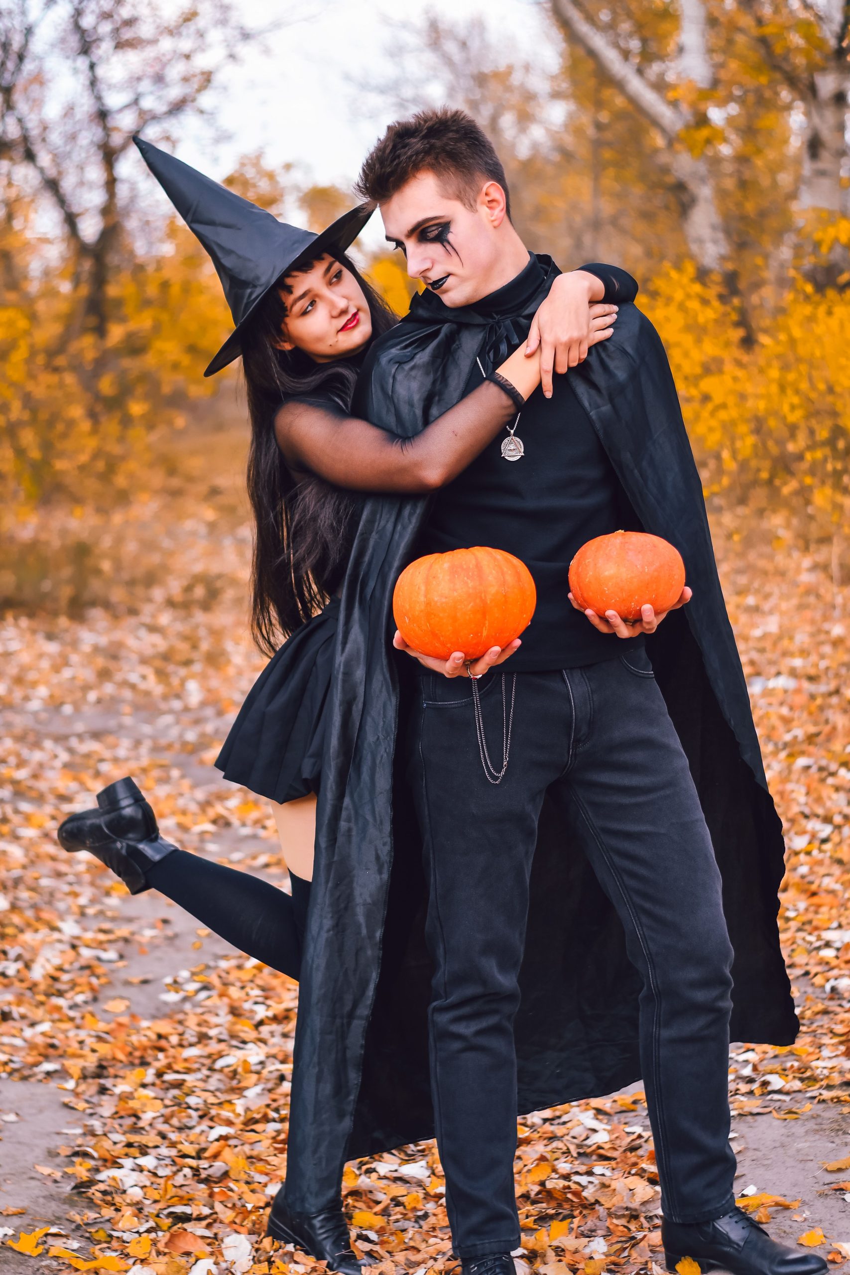 Young couple in witch and vampire costumes holding pumpkins outdoors in autumn.