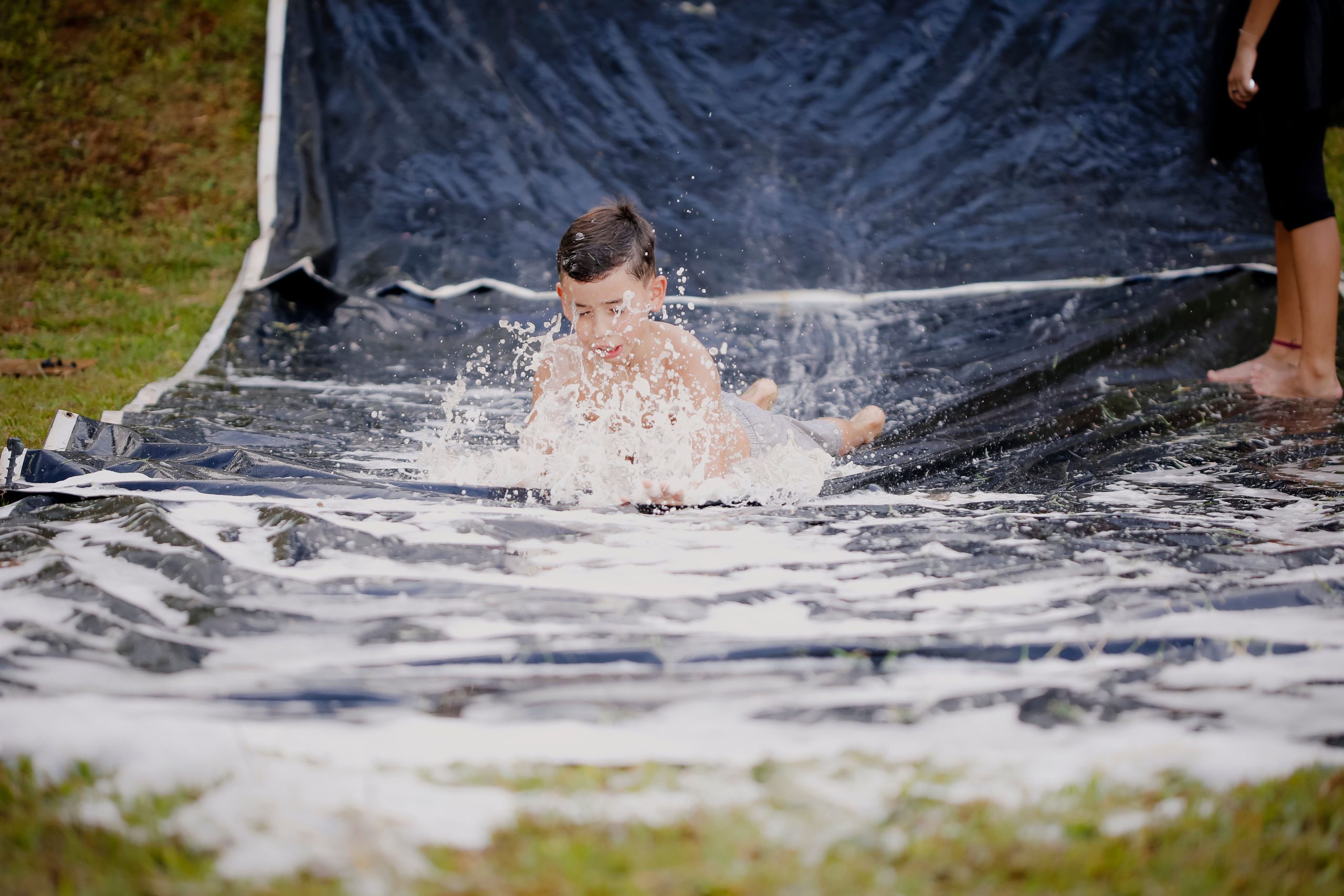 A child joyfully plays on a slip and slide outdoors, splashing water everywhere.