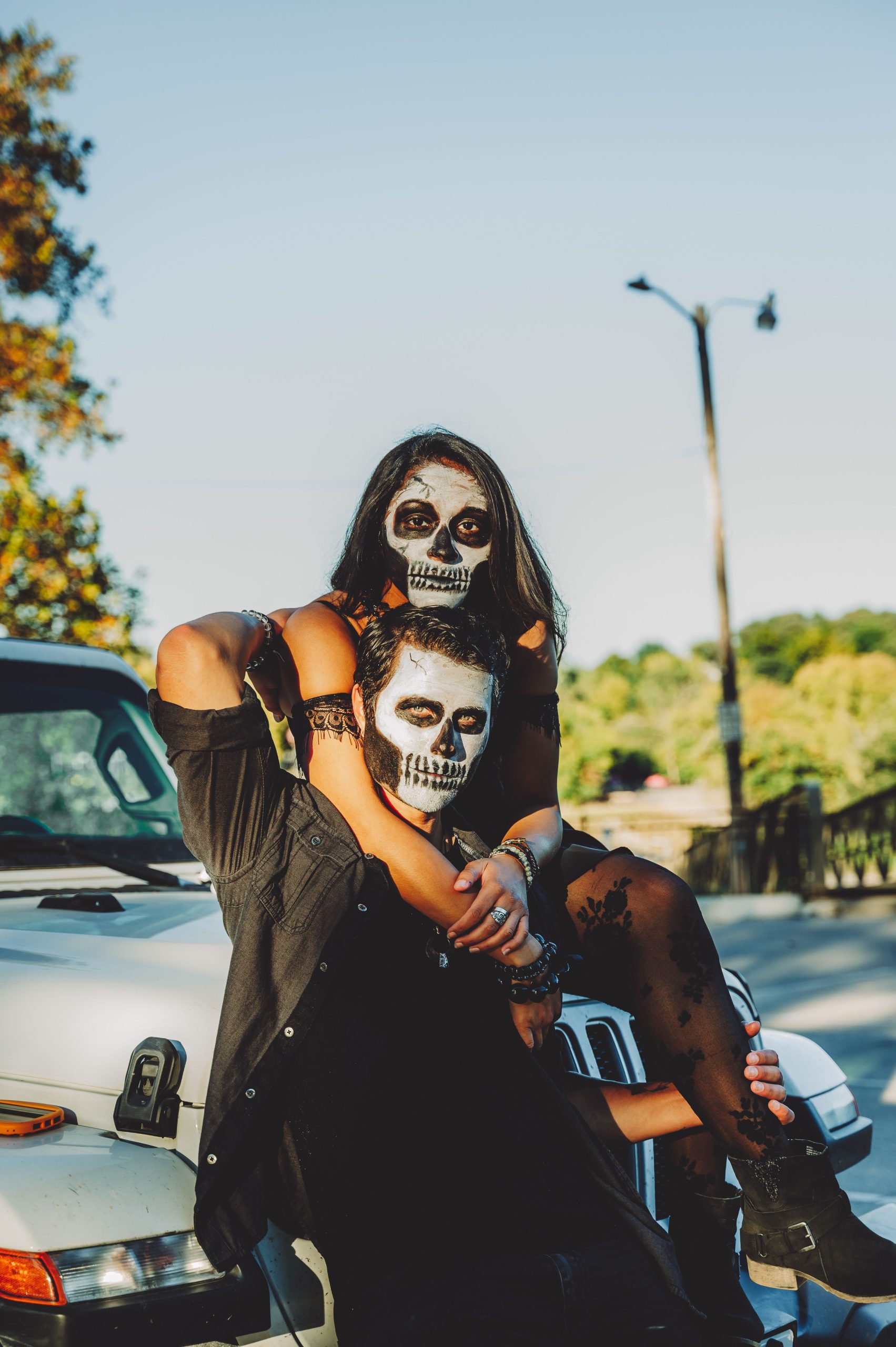 A couple with Halloween face paint sits on a car outdoors during the day.