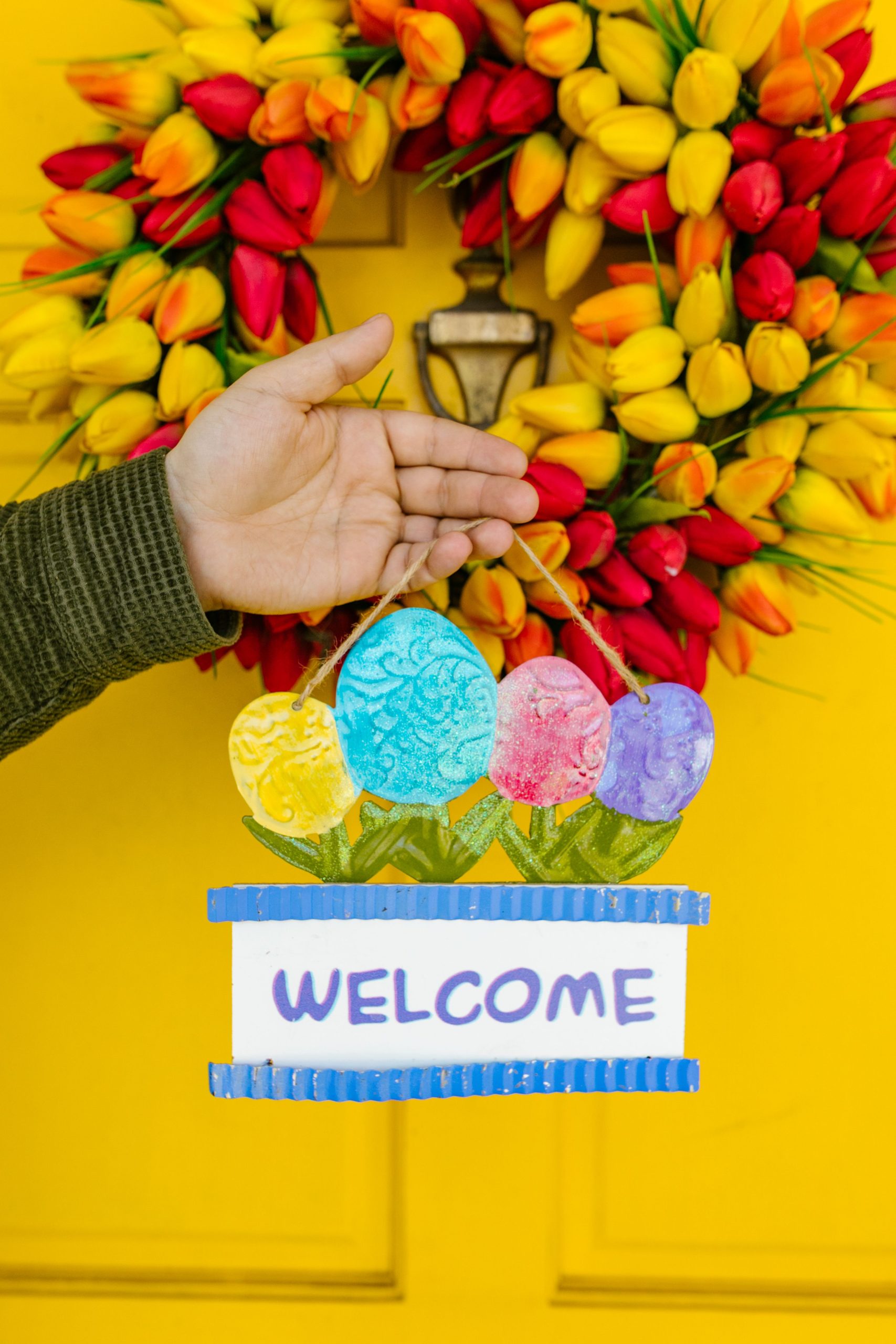 A hand holding a vibrant Easter welcome sign against a tulip wreath on a yellow door.