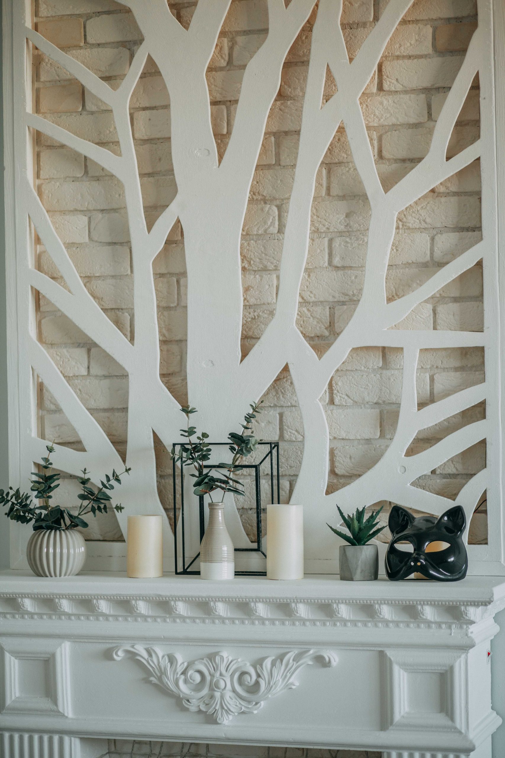 A modern mantel featuring candles, vases, and a tree wall design against a white brick background.