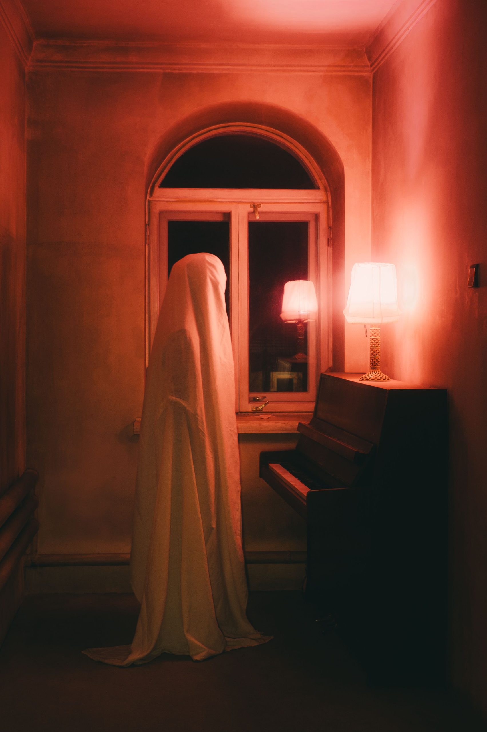 A mysterious ghostly figure stands by a piano in a dim, red-lit room, creating an eerie atmosphere.