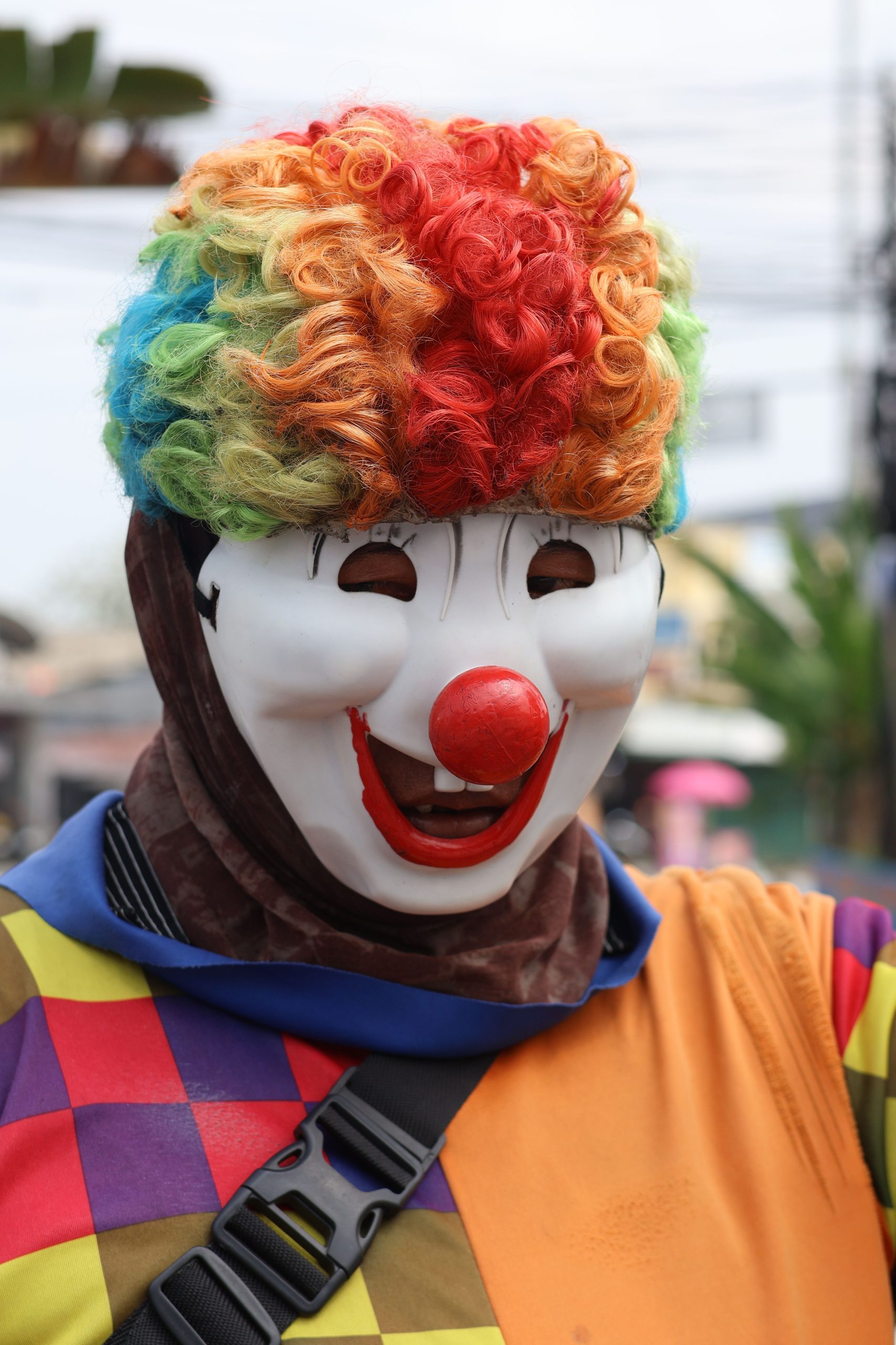 A vibrant clown in a multicolored wig and costume exudes joy in an outdoor setting.