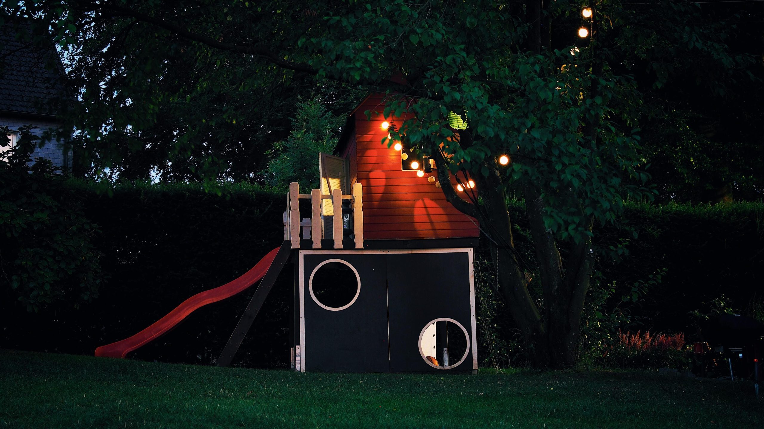 A whimsical treehouse illuminated by string lights in a lush backyard setting at dusk.