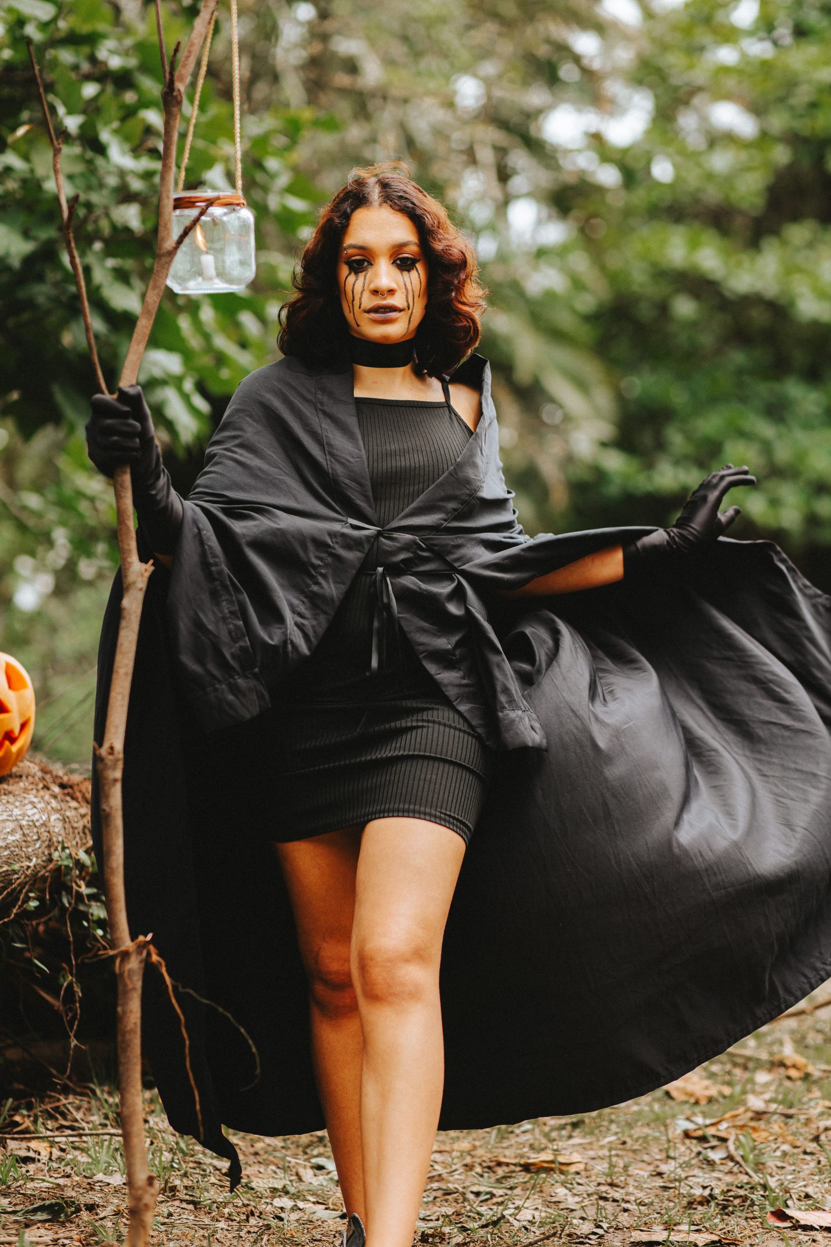 A woman dressed in a black costume carrying a lantern outdoors, creating a mysterious and Halloween vibe.