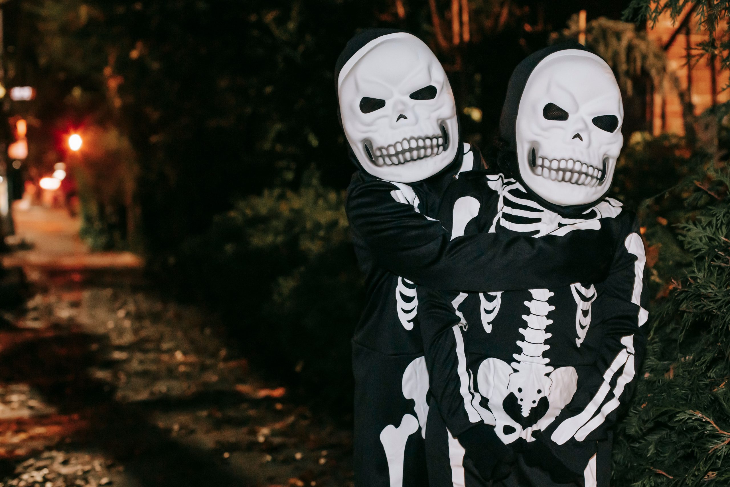 Anonymous kids in skeleton costumes and masks embracing while standing on street in evening on blurred background during Halloween celebration
