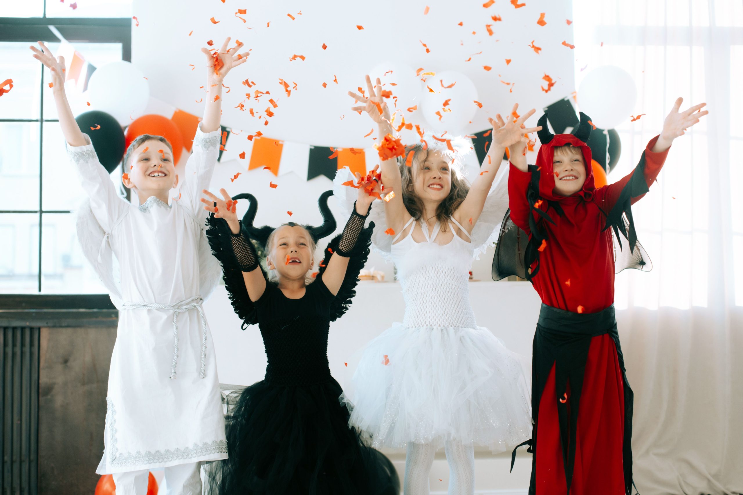 Children in angel and demon costumes celebrating Halloween indoors with confetti and balloons.