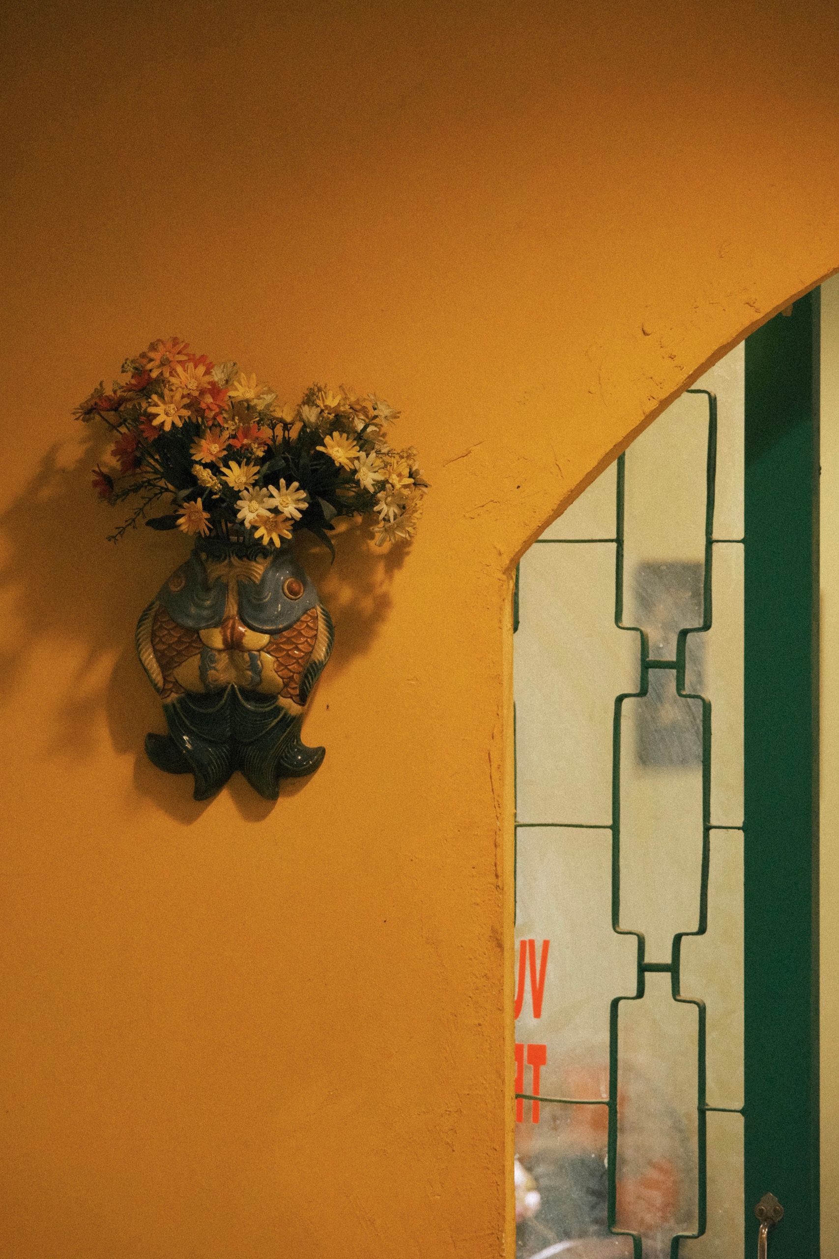 Decorative fish vase with flowers on a vibrant yellow wall, next to a stained glass window.