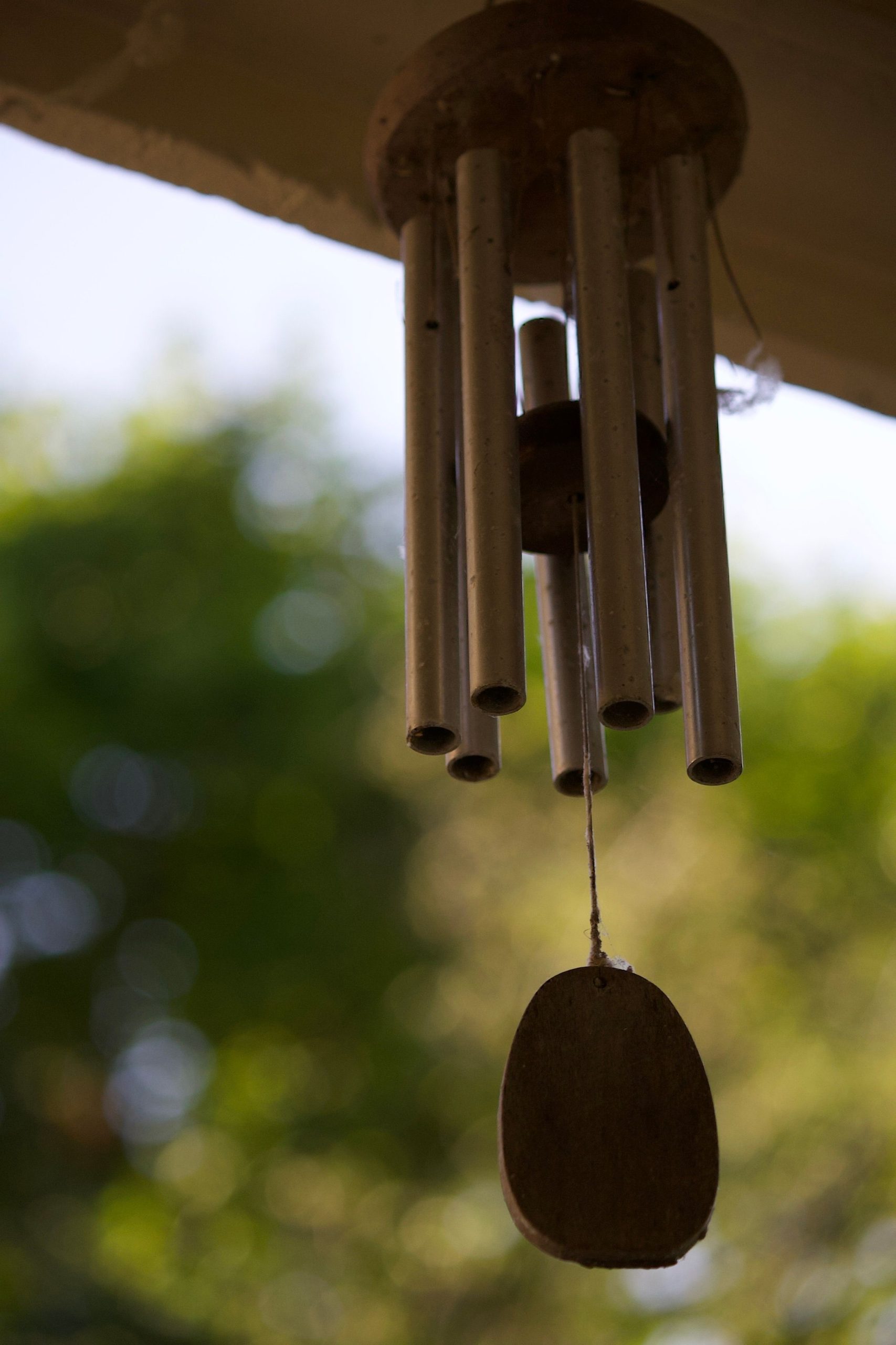 Elegant metal wind chime hanging outdoors with sunlit trees in the background.