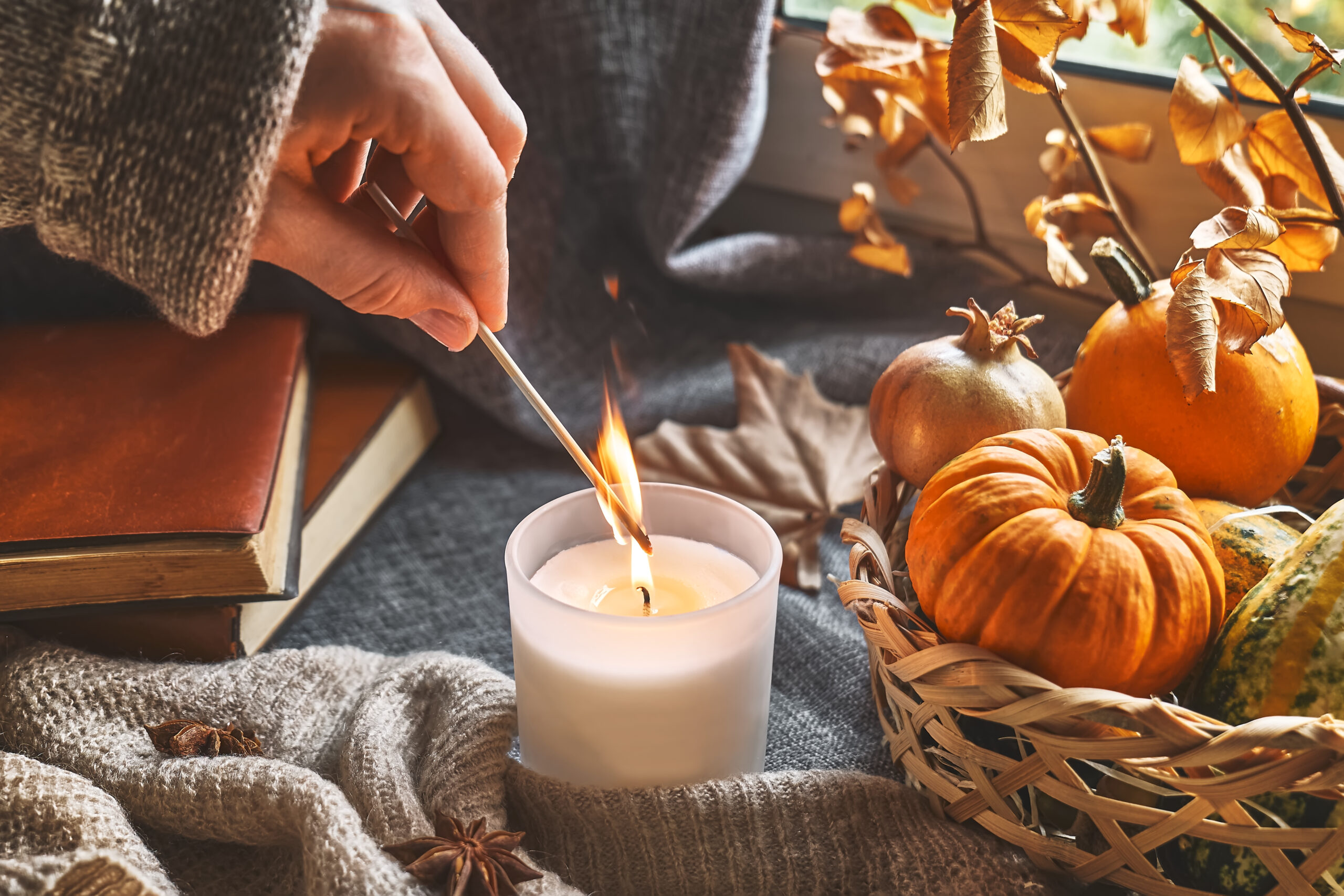 Hand with burning match lighting a candle on the windowsill with cozy autumn still life with pumpkins, knitted woolen sweater and books. Autumn home decor. Cozy fall mood. Thanksgiving. Halloween. -