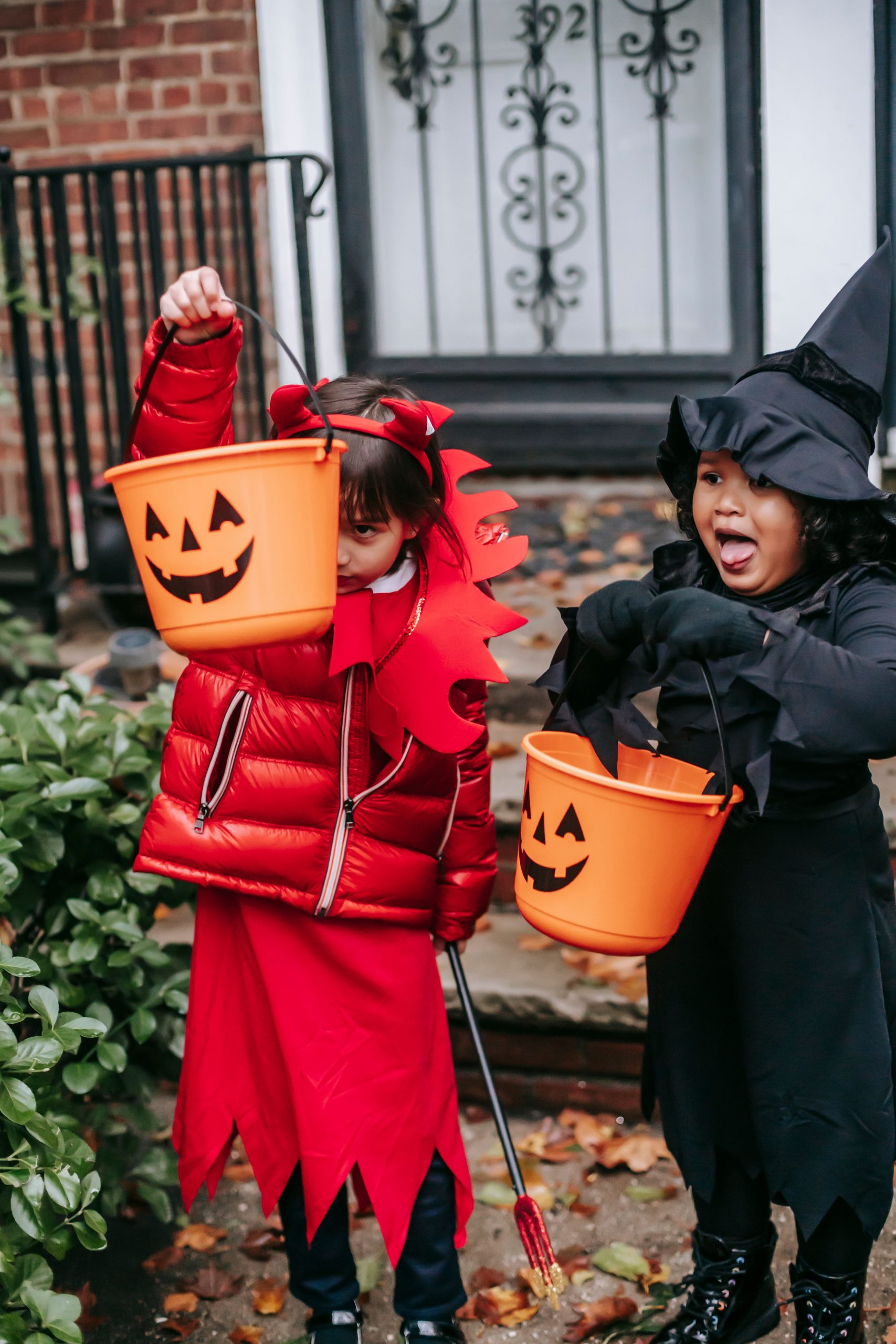 Little girls in red devil costume with horns and pitchfork and in black witch costume with hat with orange buckets on Halloween standing near house door in autumn