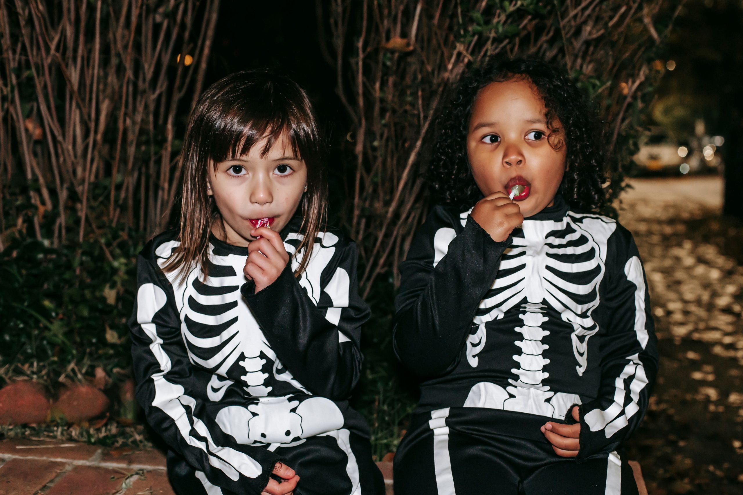 Positive little multiracial girls wearing traditional skeleton costumes and enjoying sweet stick candies while sitting on brick border in autumn park after playing Trick or Treat game