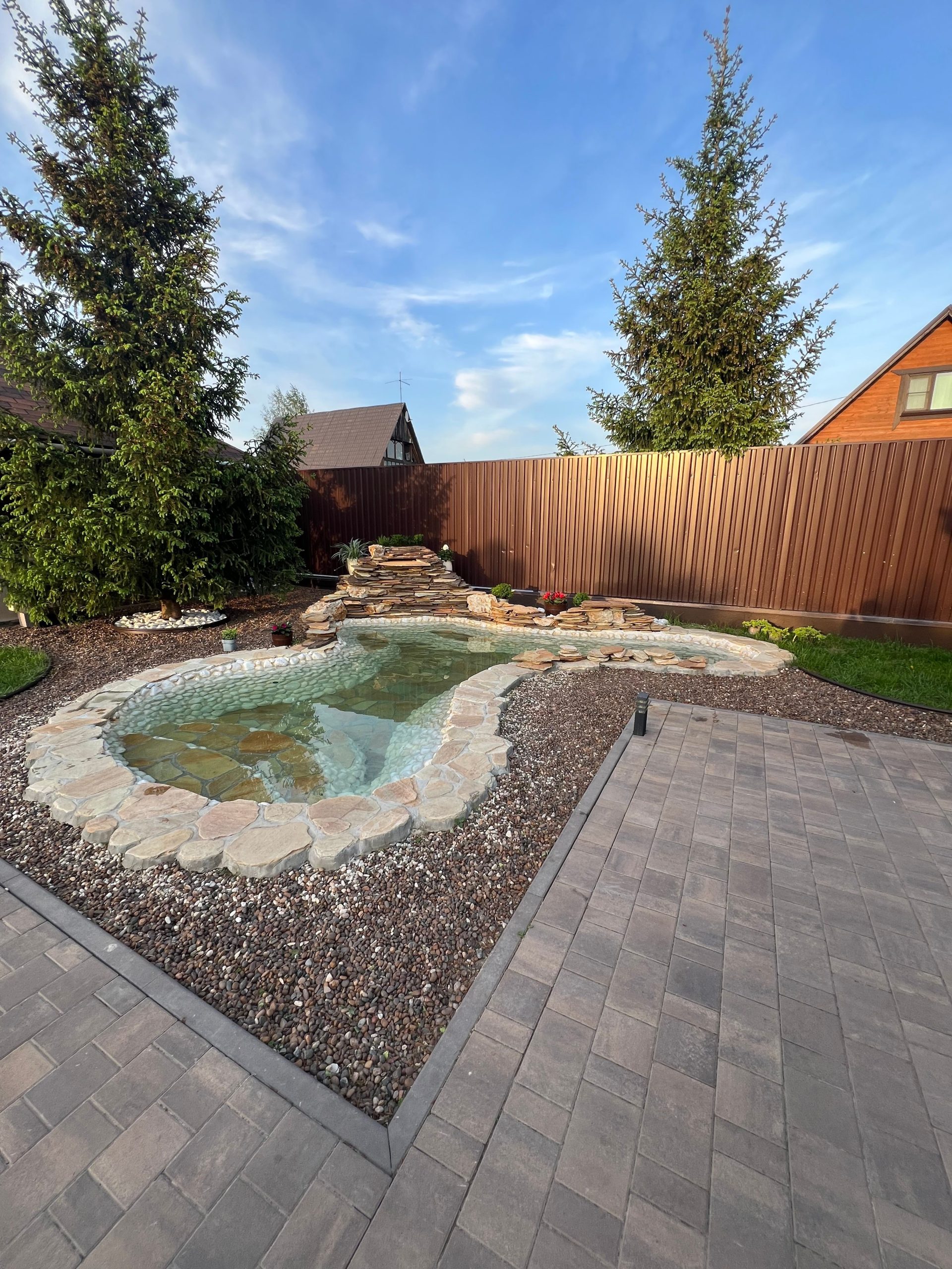 Serene backyard with stone pond, trees, and patio under a blue sky.