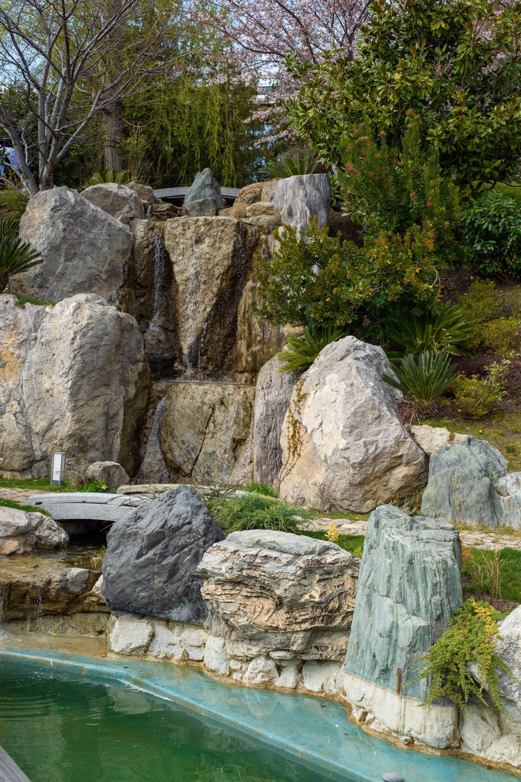Tranquil scene of a cascading waterfall over natural rocks surrounded by greenery in a garden setting.