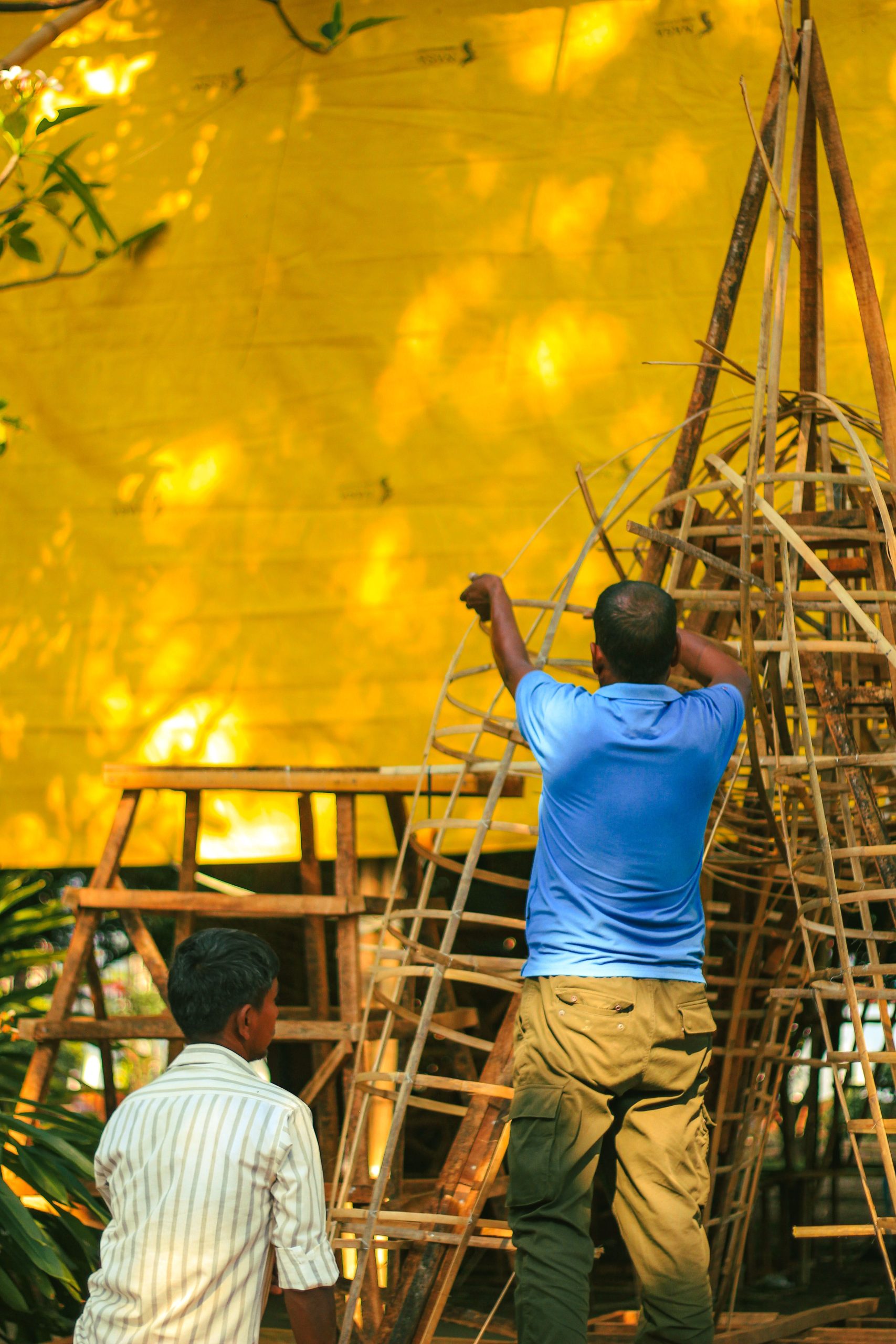 Two artisans constructing a bamboo framework against a vibrant yellow backdrop outdoors.