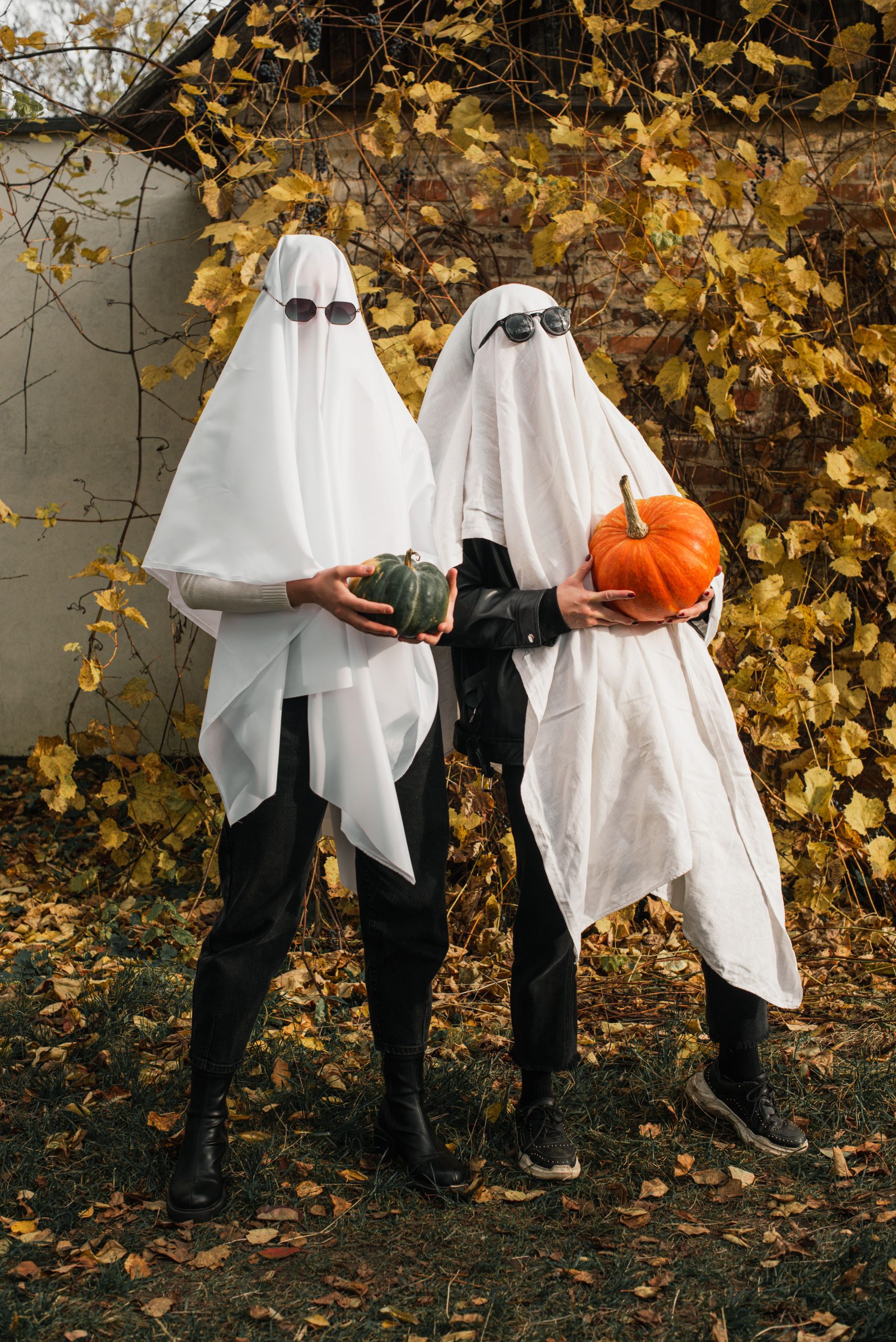 Two people dressed as ghosts holding pumpkins against autumn backdrop.