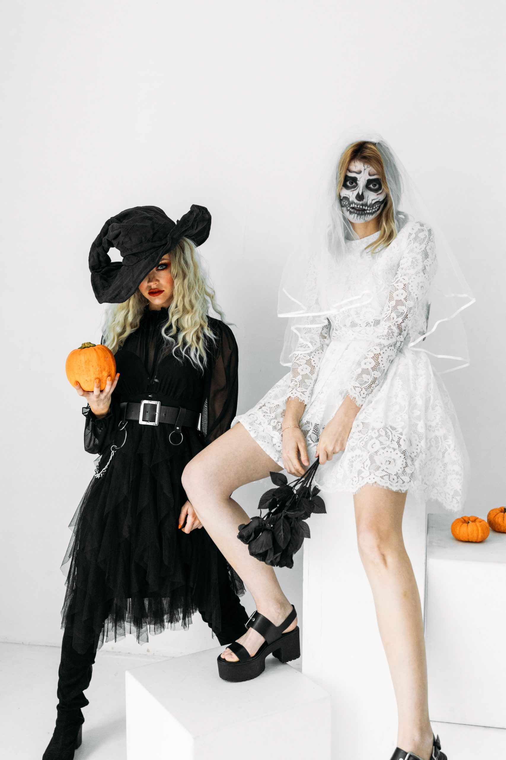 Two women in Halloween costumes: a witch holding a pumpkin and a skeleton bride with face paint.
