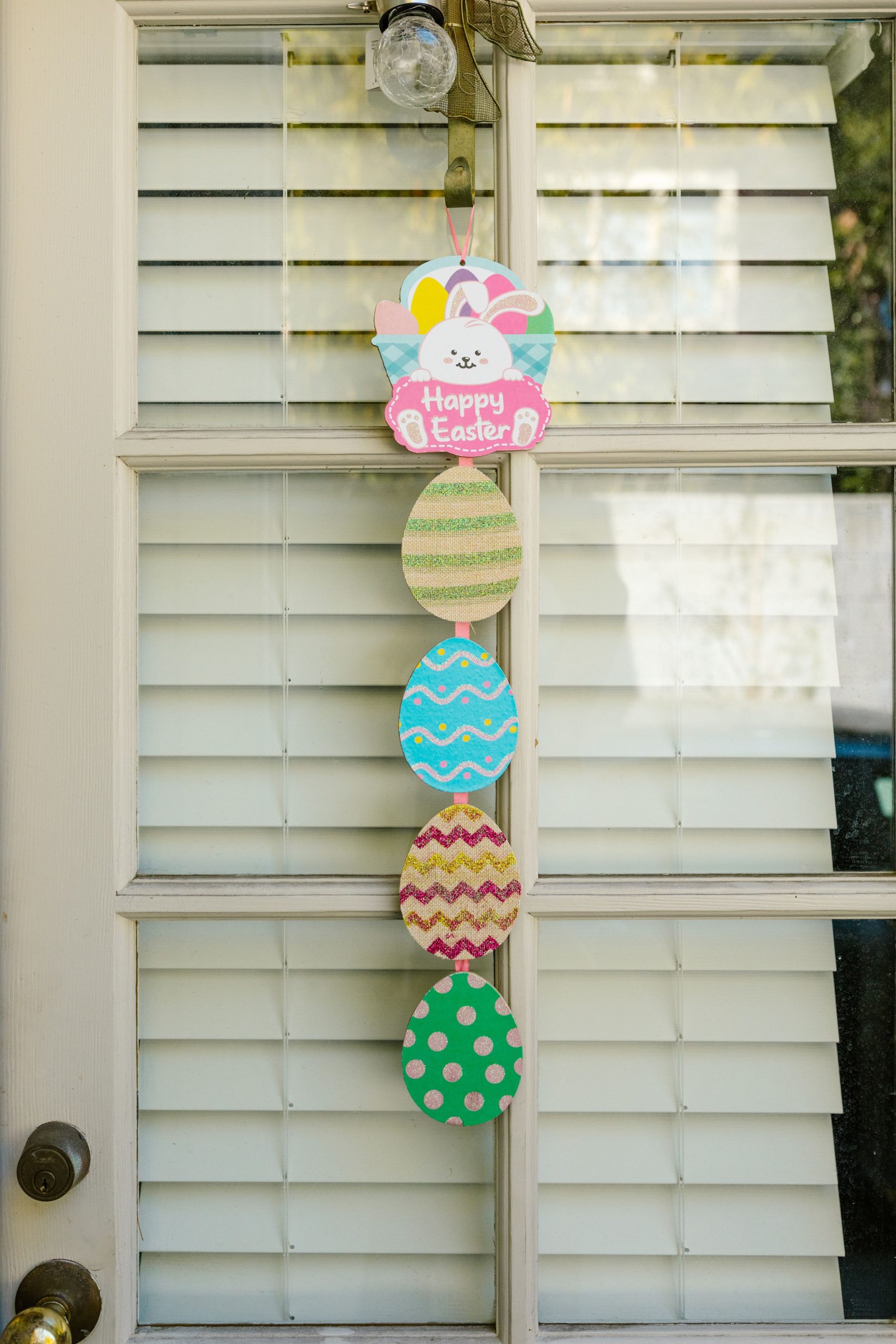 Vibrant Easter eggs and bunny decor hanging on glass door with window blinds.