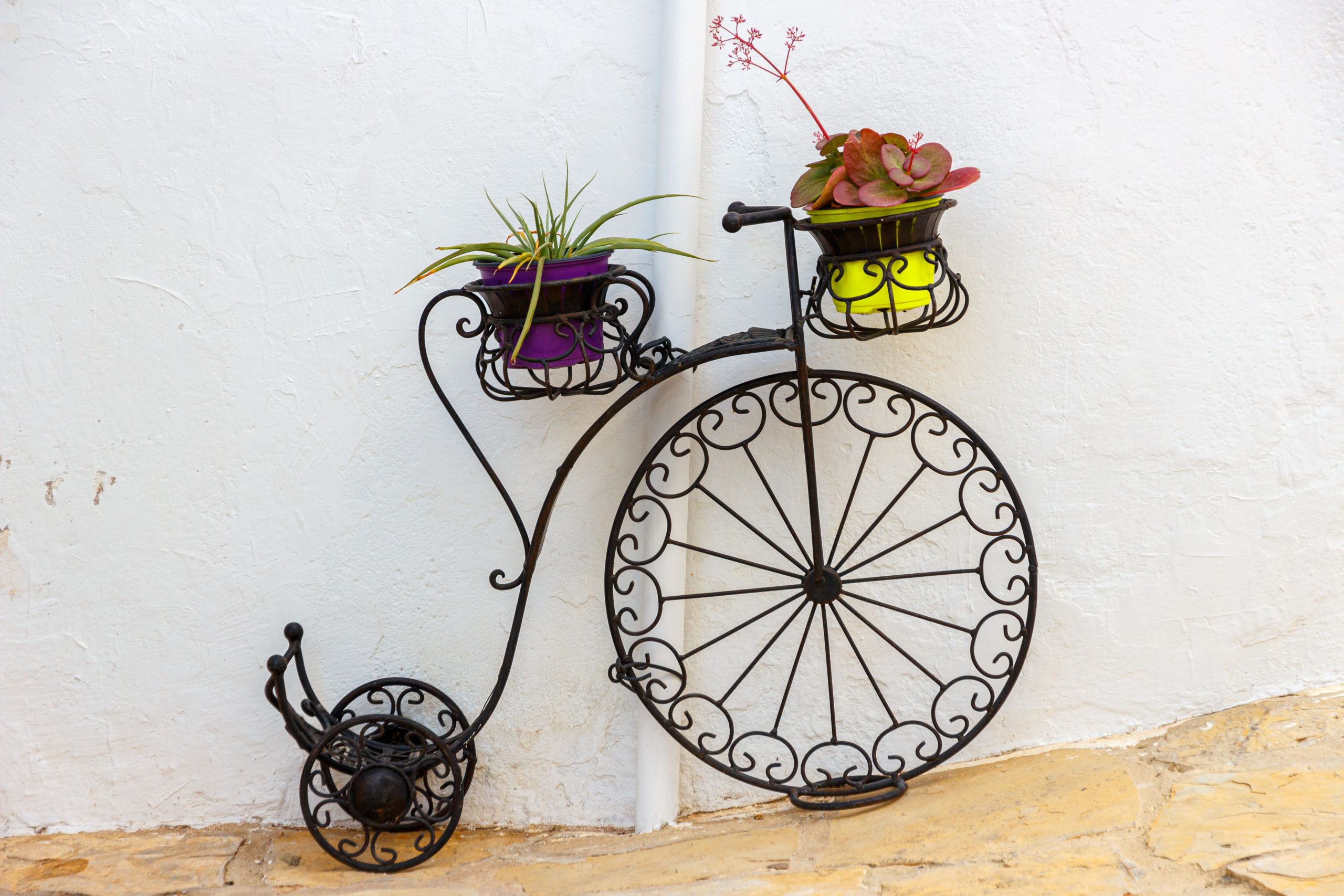 Vintage wrought iron bicycle planter with vibrant potted plants against a clean white wall for outdoor decor.