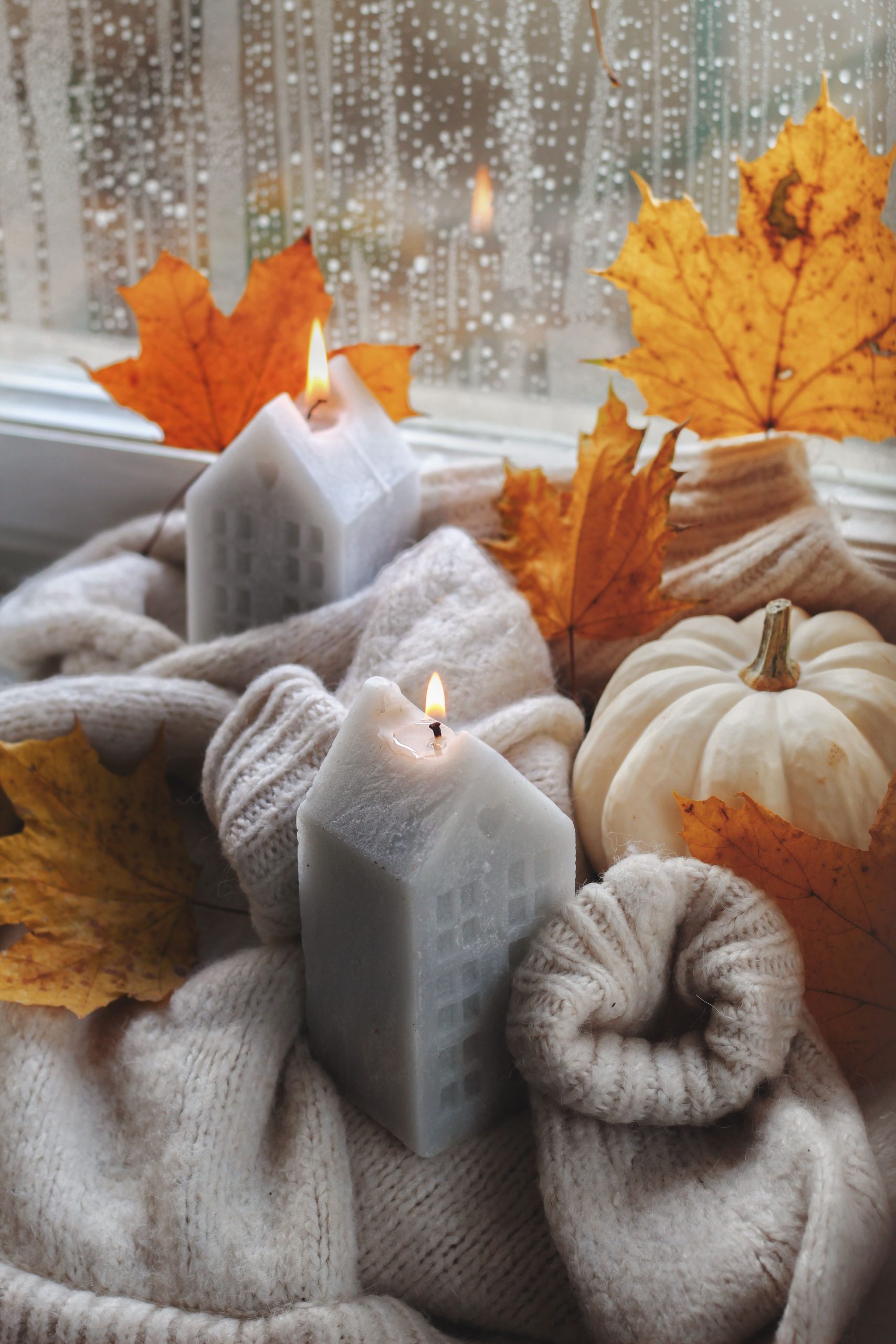 Warm autumn scene with glowing candles, knitwear, and leaves by the window.