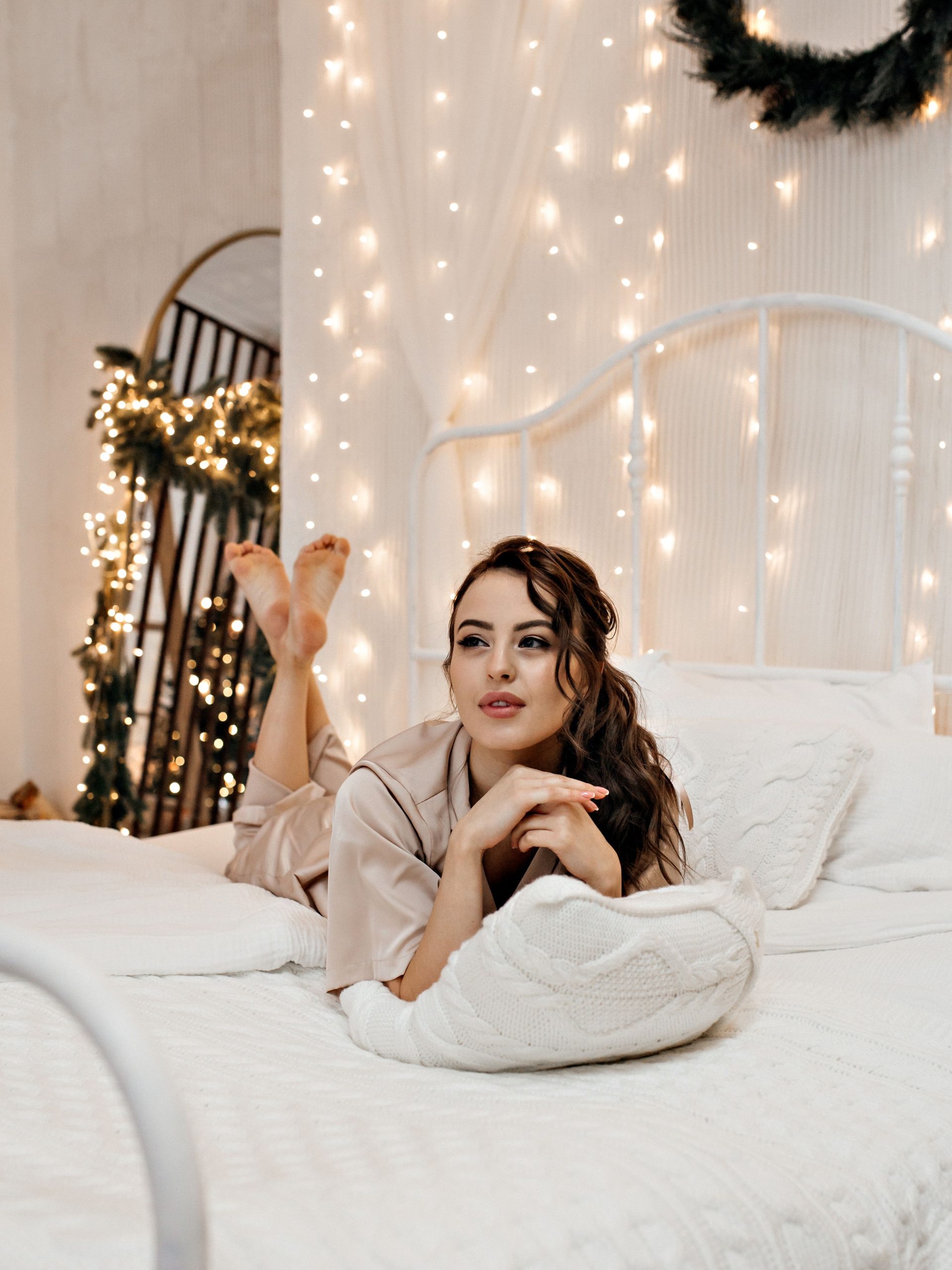 Woman relaxing on bed in pajamas with festive lights and decor.