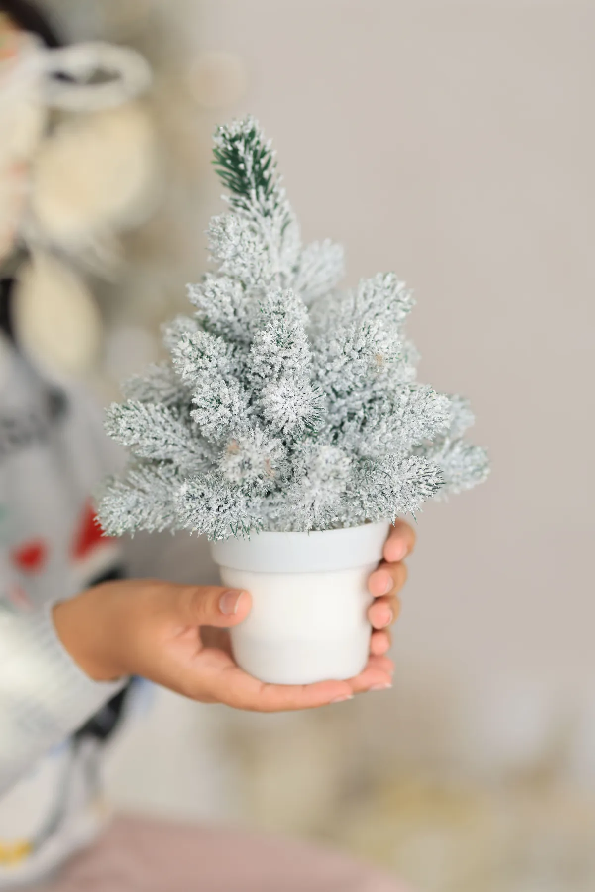 A child's hands holding a frosted mini Christmas tree in a white pot, perfect for holiday decor.