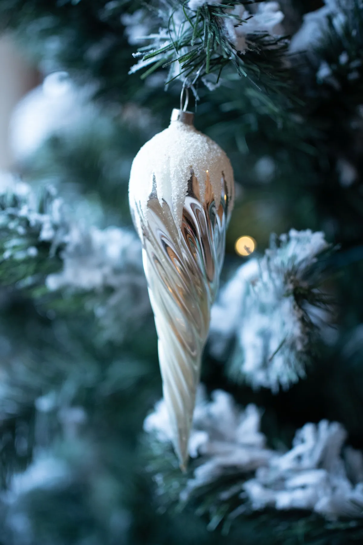 A close-up of a shimmering silver Christmas ornament hanging from a snow-covered evergreen branch.