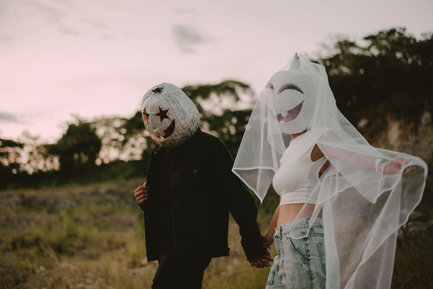 A couple wearing pumpkin head costumes and veils walking hand in hand in a rural setting.