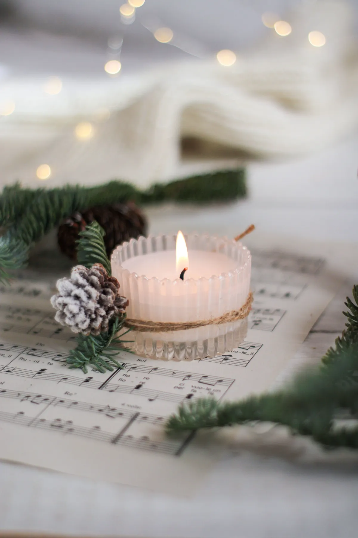 A lit candle with spruce twigs and pinecones rests on a musical sheet, creating warm winter ambiance.