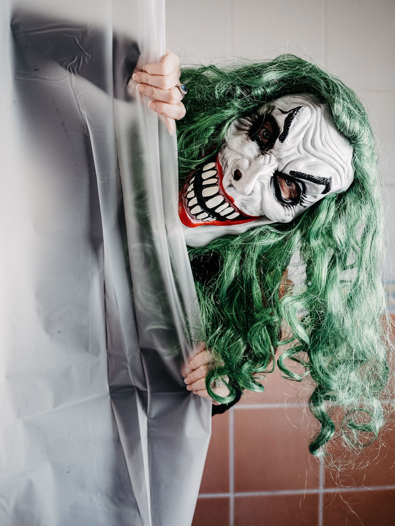A spooky figure with a joker mask and green hair peeks from behind a shower curtain.