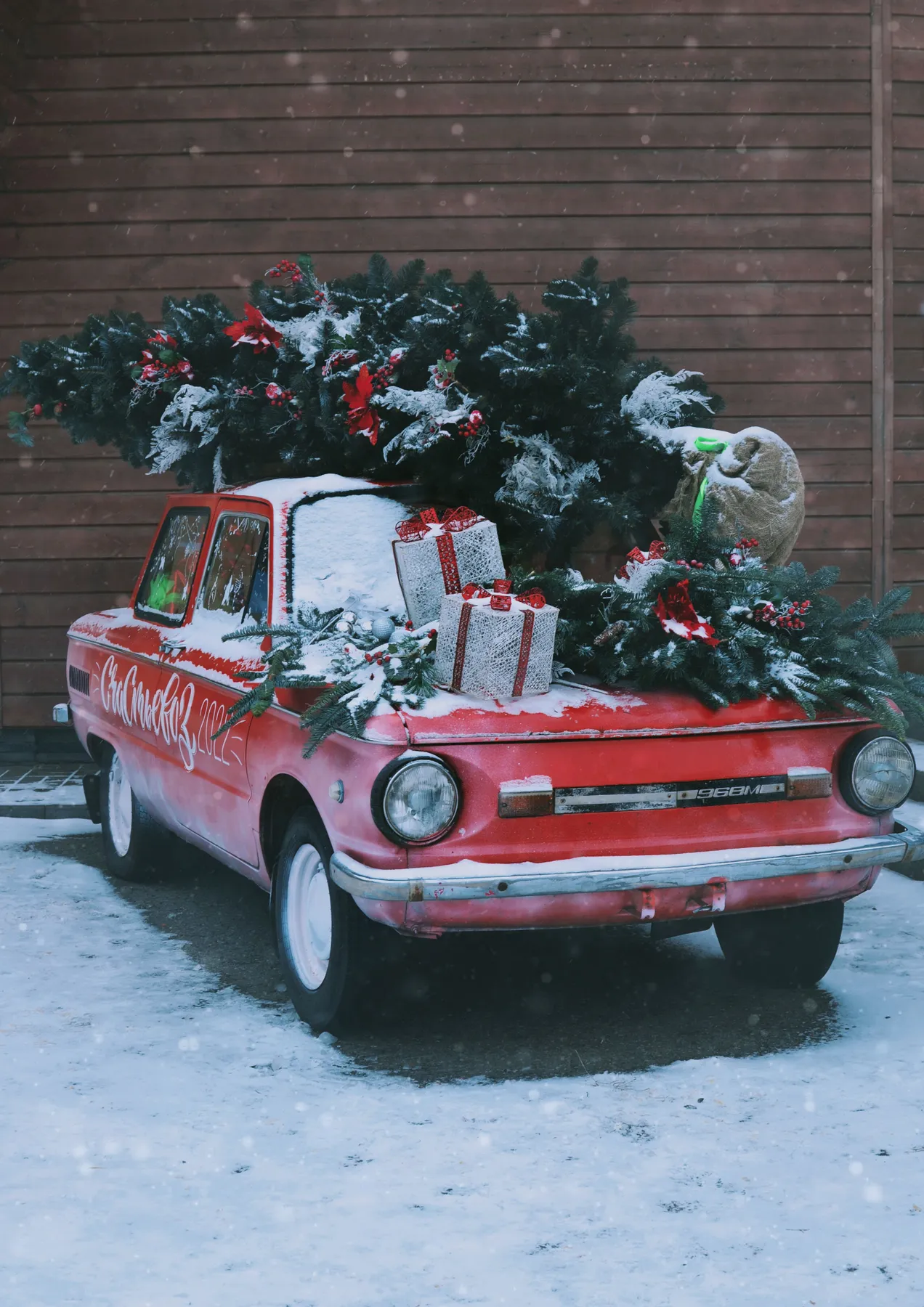A vintage car covered in snow, adorned with a Christmas tree and gifts, captures the holiday spirit.