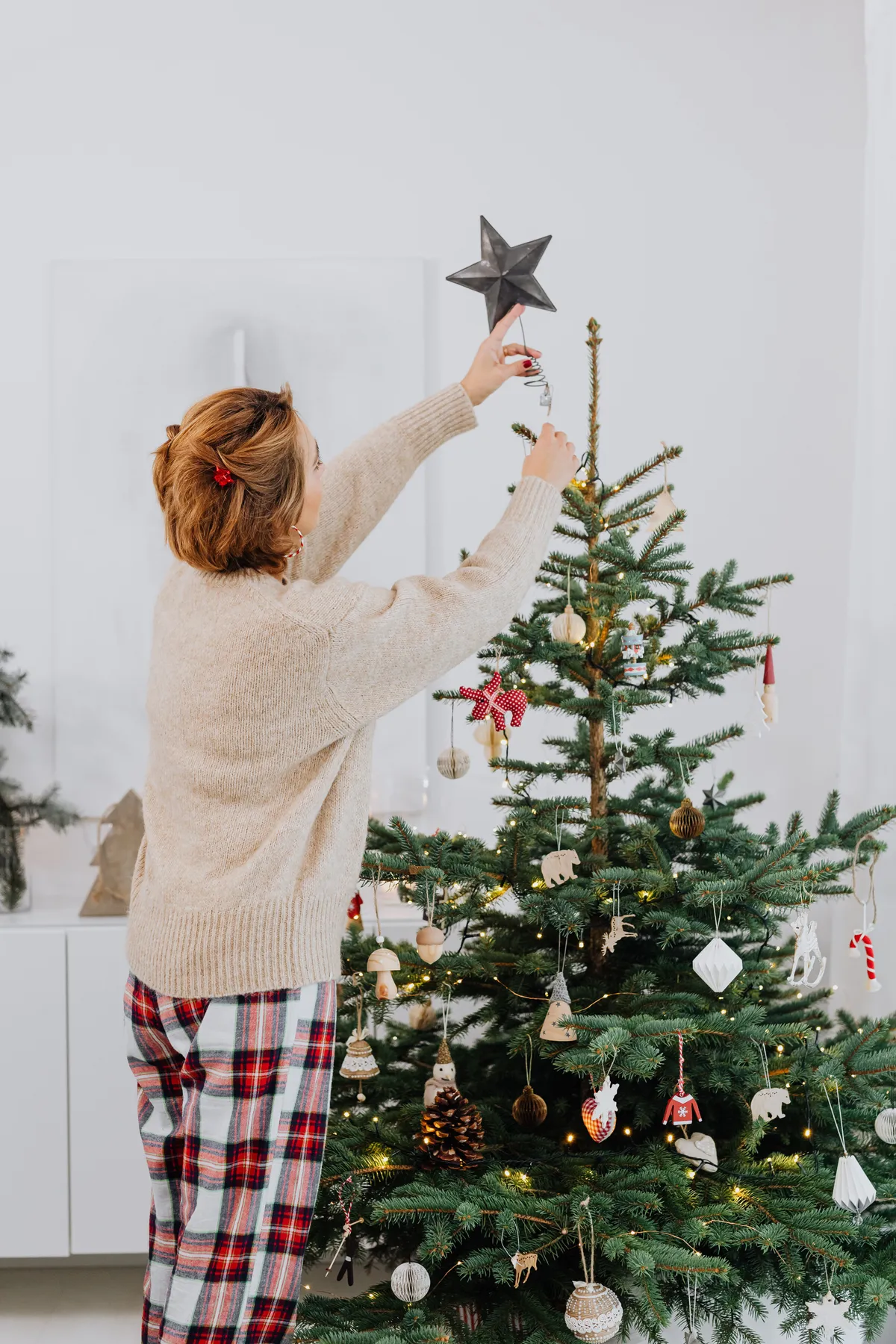 A woman in cozy attire places a star on a Christmas tree, capturing the festive spirit indoors.
