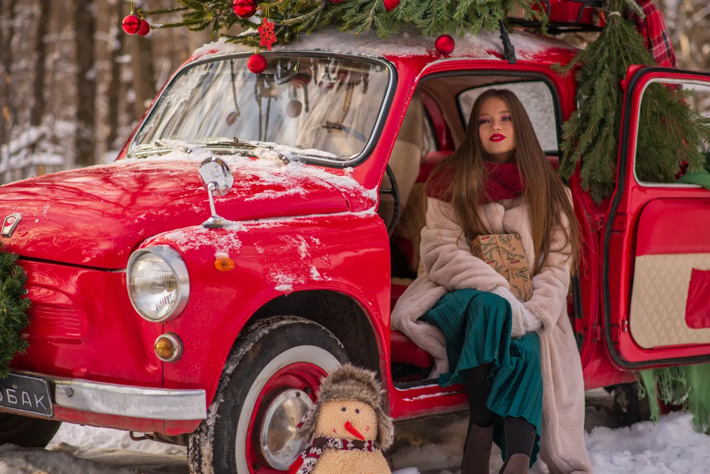 A woman in warm clothing sits by a Christmas-themed car in a snowy forest.