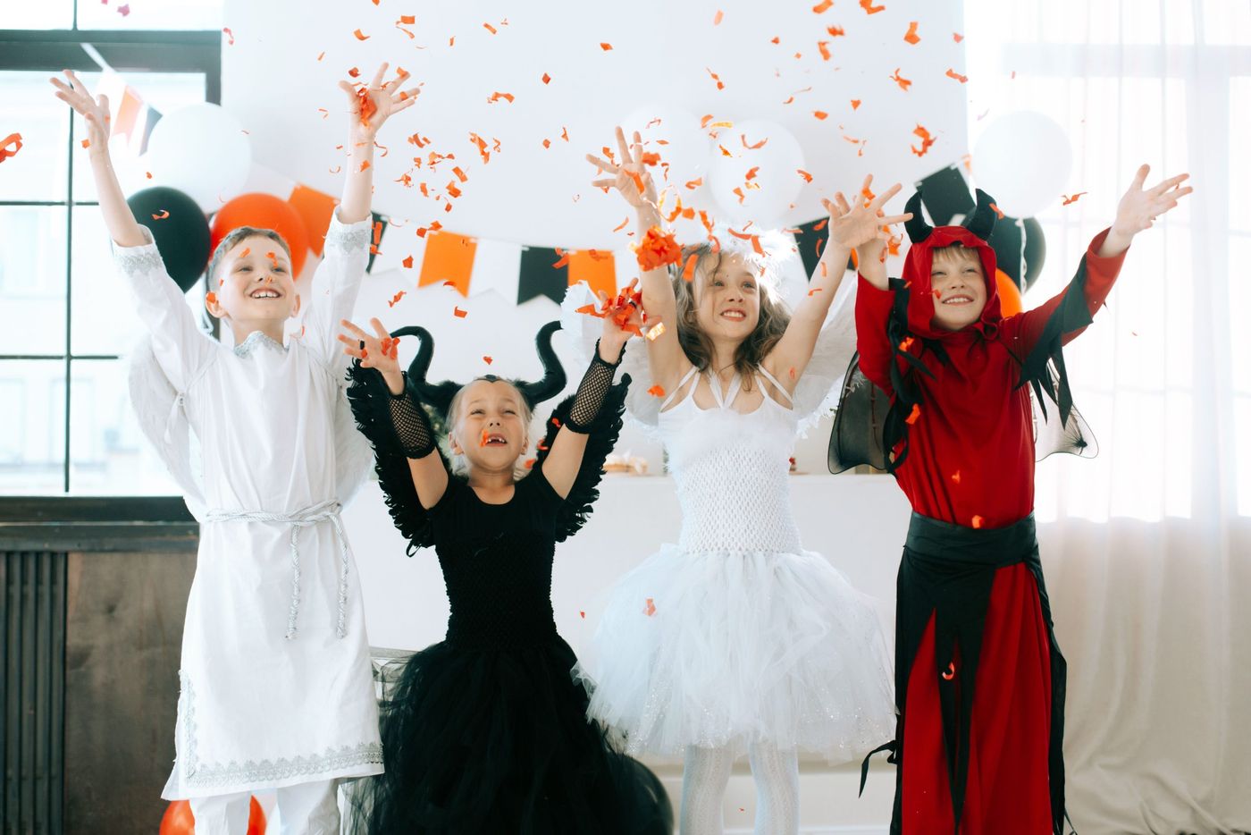 Children in angel and demon costumes celebrating Halloween indoors with confetti and balloons.