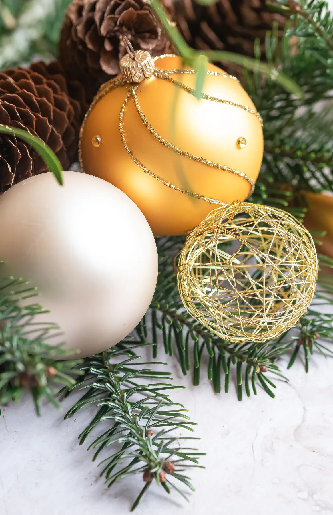Close-up of gold and white Xmas ornaments amidst pine needles and cones.