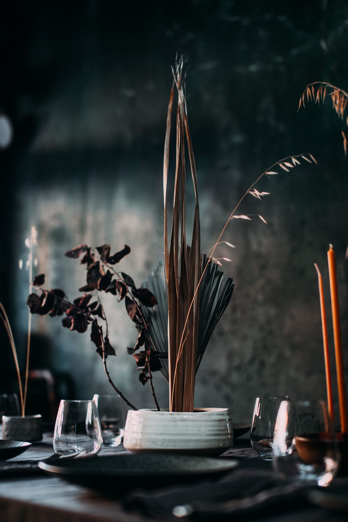 Composition of dry leaves and branches in white vase placed on table with dark tablecloth and utensil among candles and glasses with dark wall on background