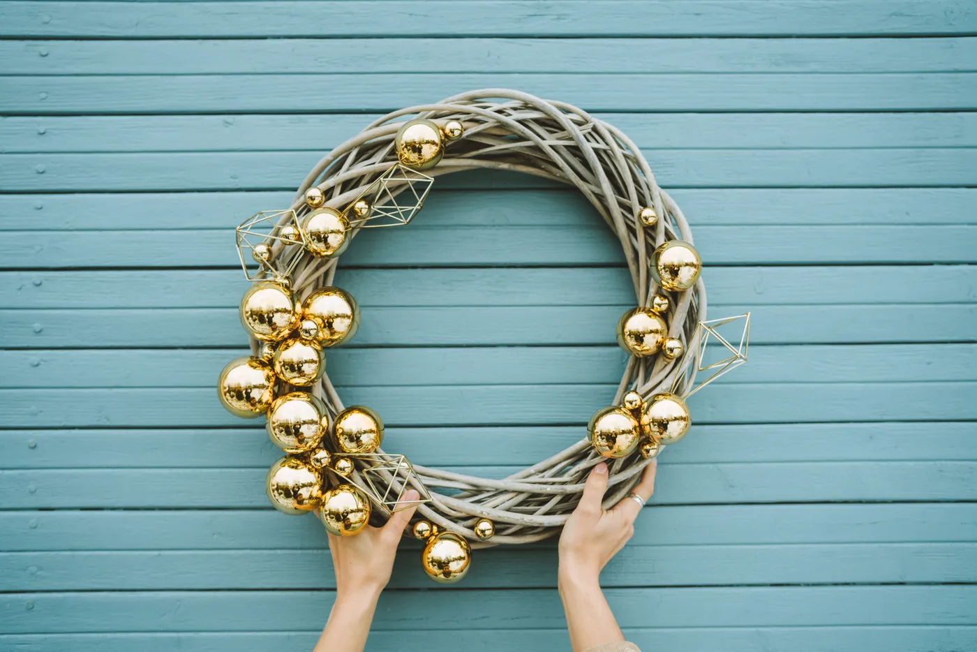 Hand holding an elegant Christmas wreath decorated with golden ornaments against a blue wooden background.