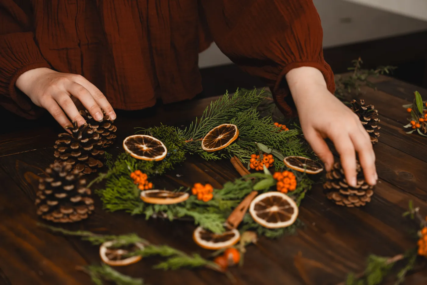 Hands arranging pine cones and dried oranges on a festive holiday wreath.