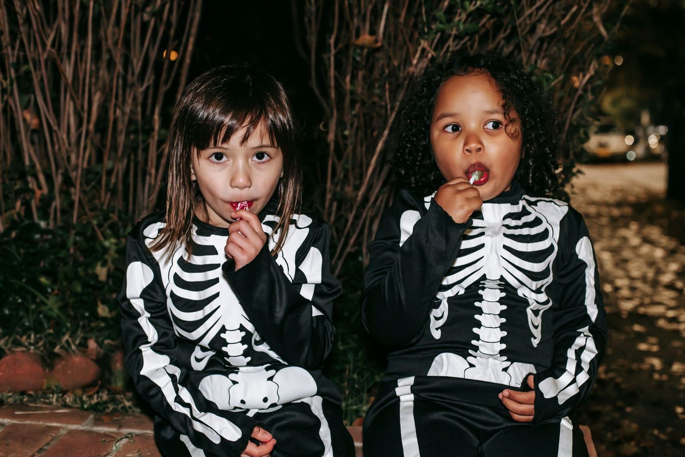 Positive little multiracial girls wearing traditional skeleton costumes and enjoying sweet stick candies while sitting on brick border in autumn park after playing Trick or Treat game