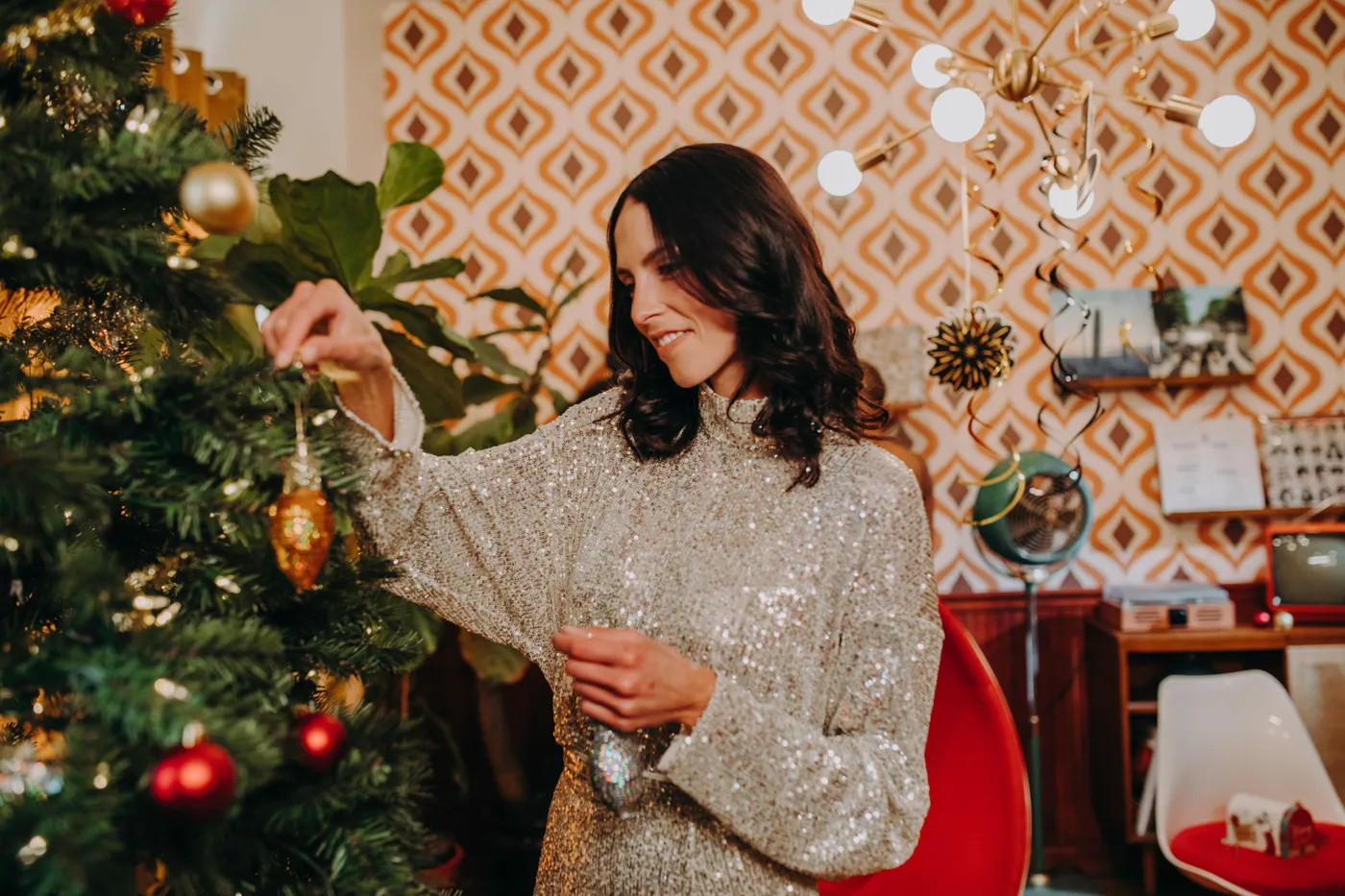 Smiling woman in a sparkling dress decorates a Christmas tree indoors, creating a festive holiday atmosphere.