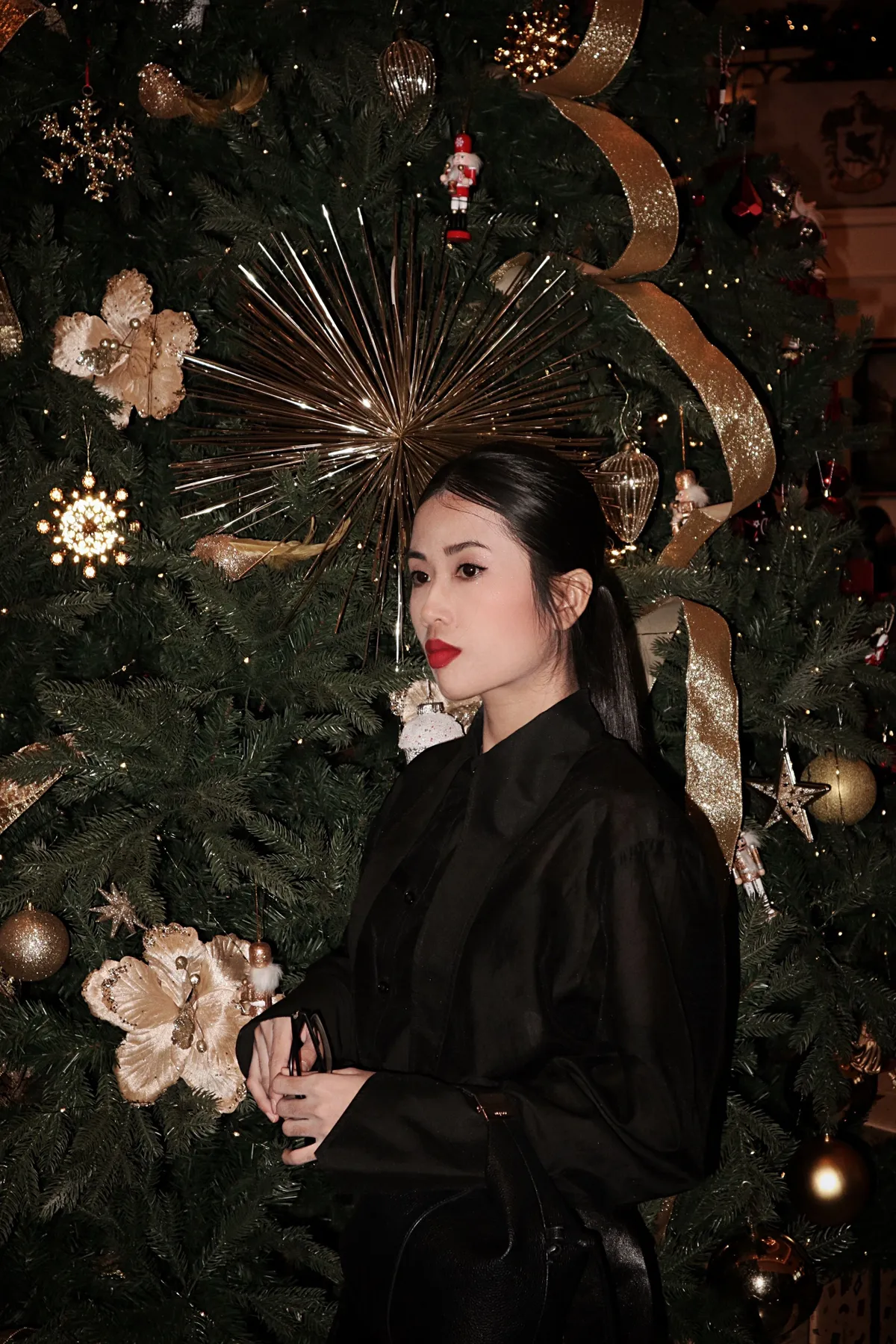 Stylish woman in black beside a festive Christmas tree adorned with gold ornaments.